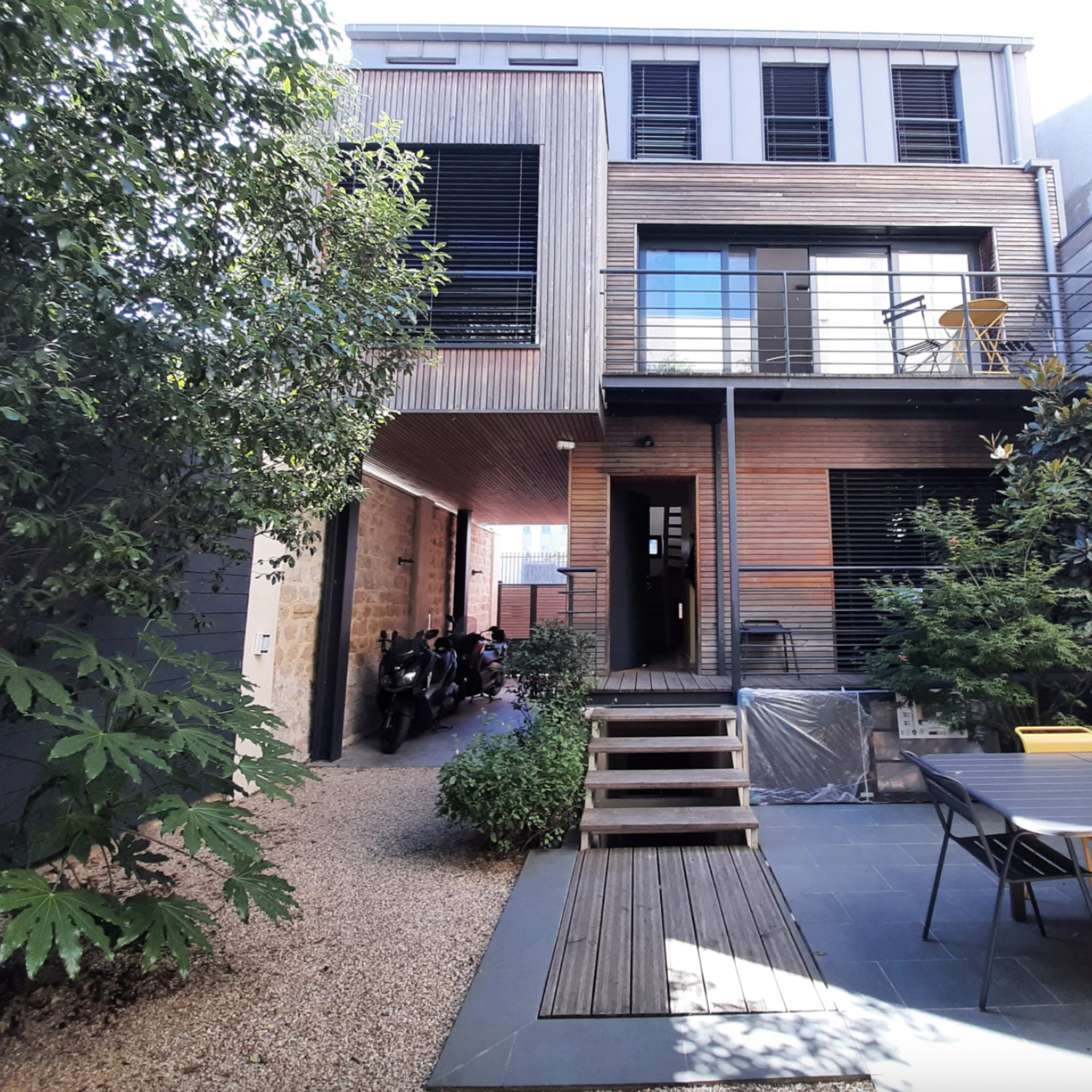 A modern house with a wooden exterior, a patio area, and a garden surrounded by plants and gravel pathways.
