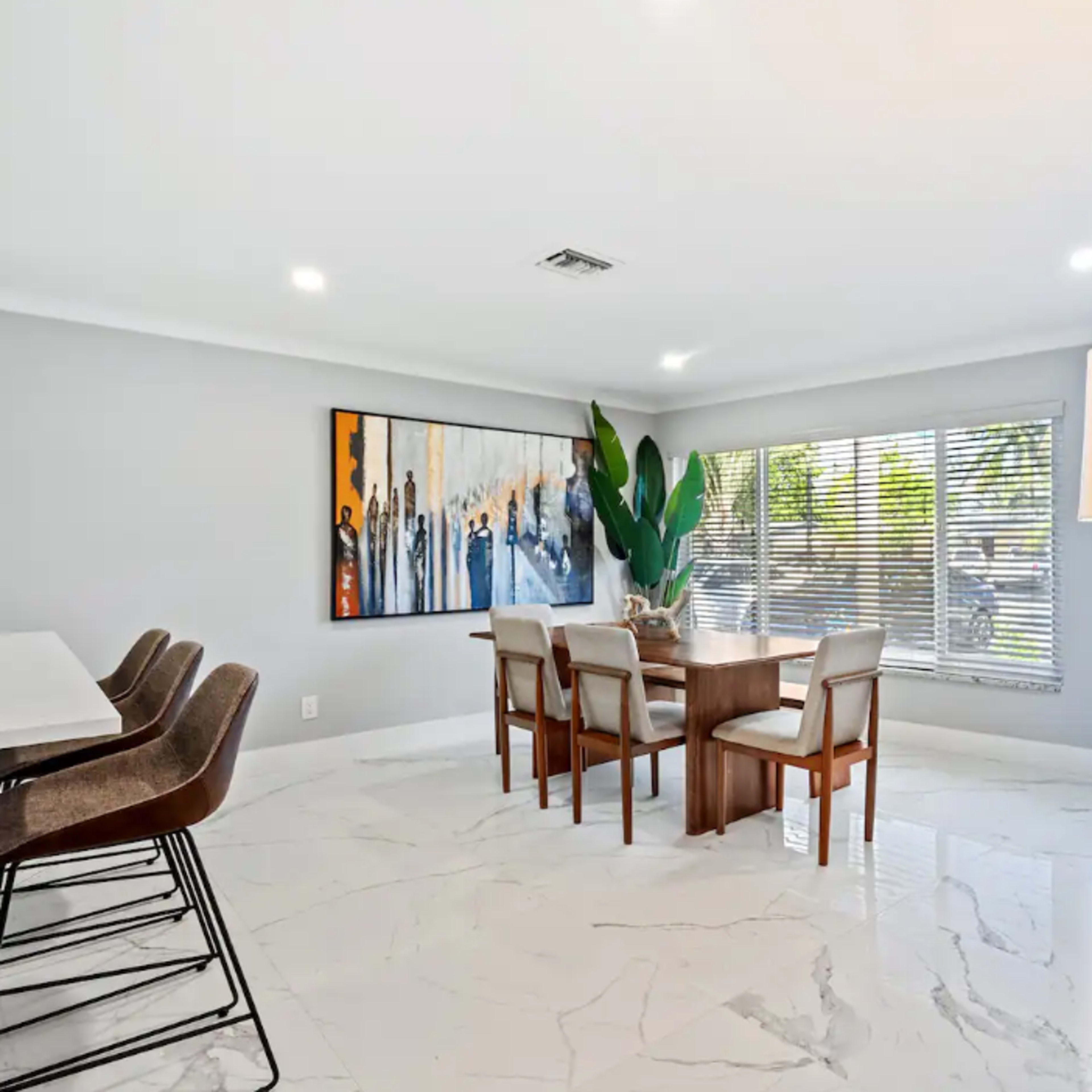 A modern dining area features a wooden dining table surrounded by chairs, with a large painting on the wall and a lamp on a side table.