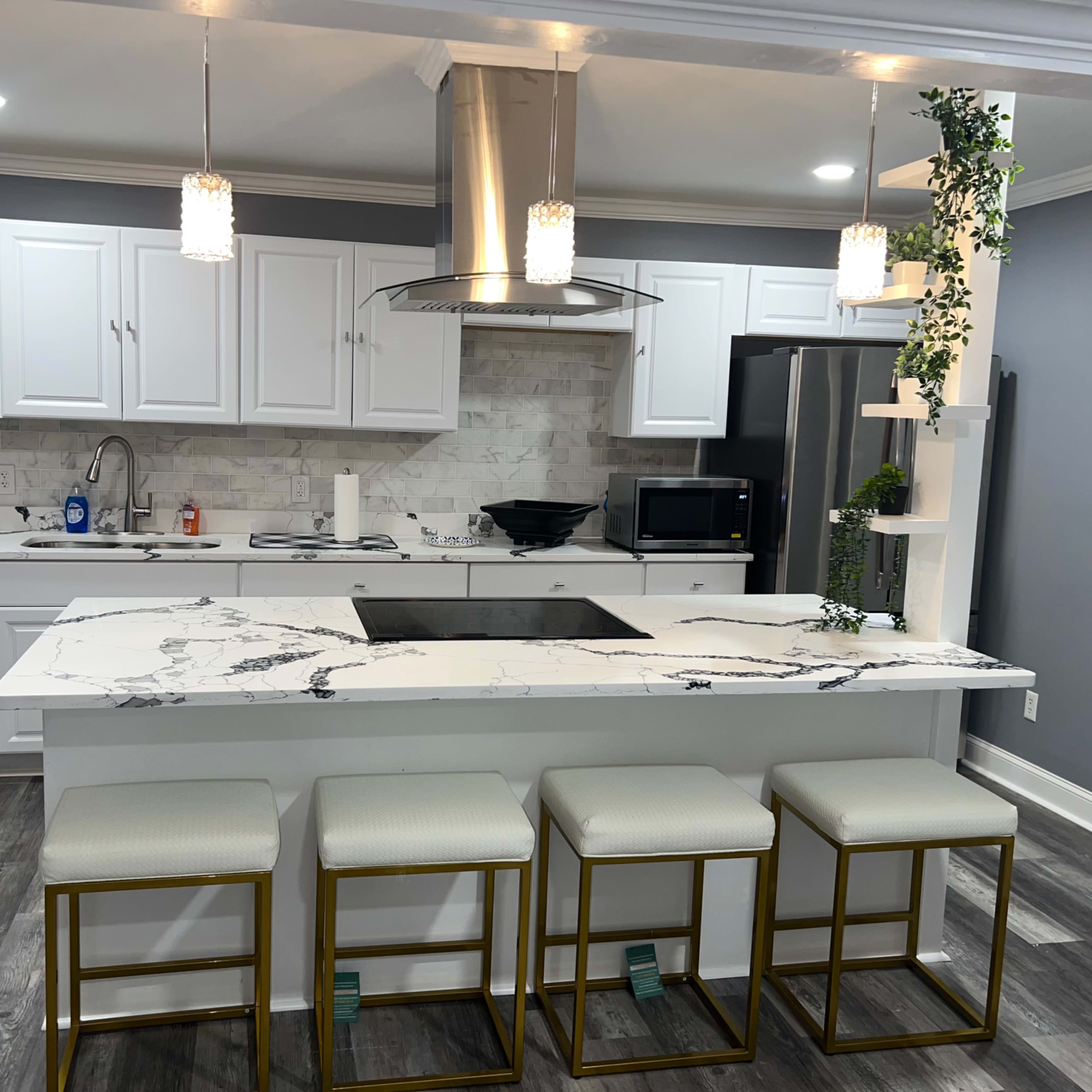 A modern kitchen features white cabinetry, a marble countertop with an integrated cooktop, and four gold-framed stools at the island.