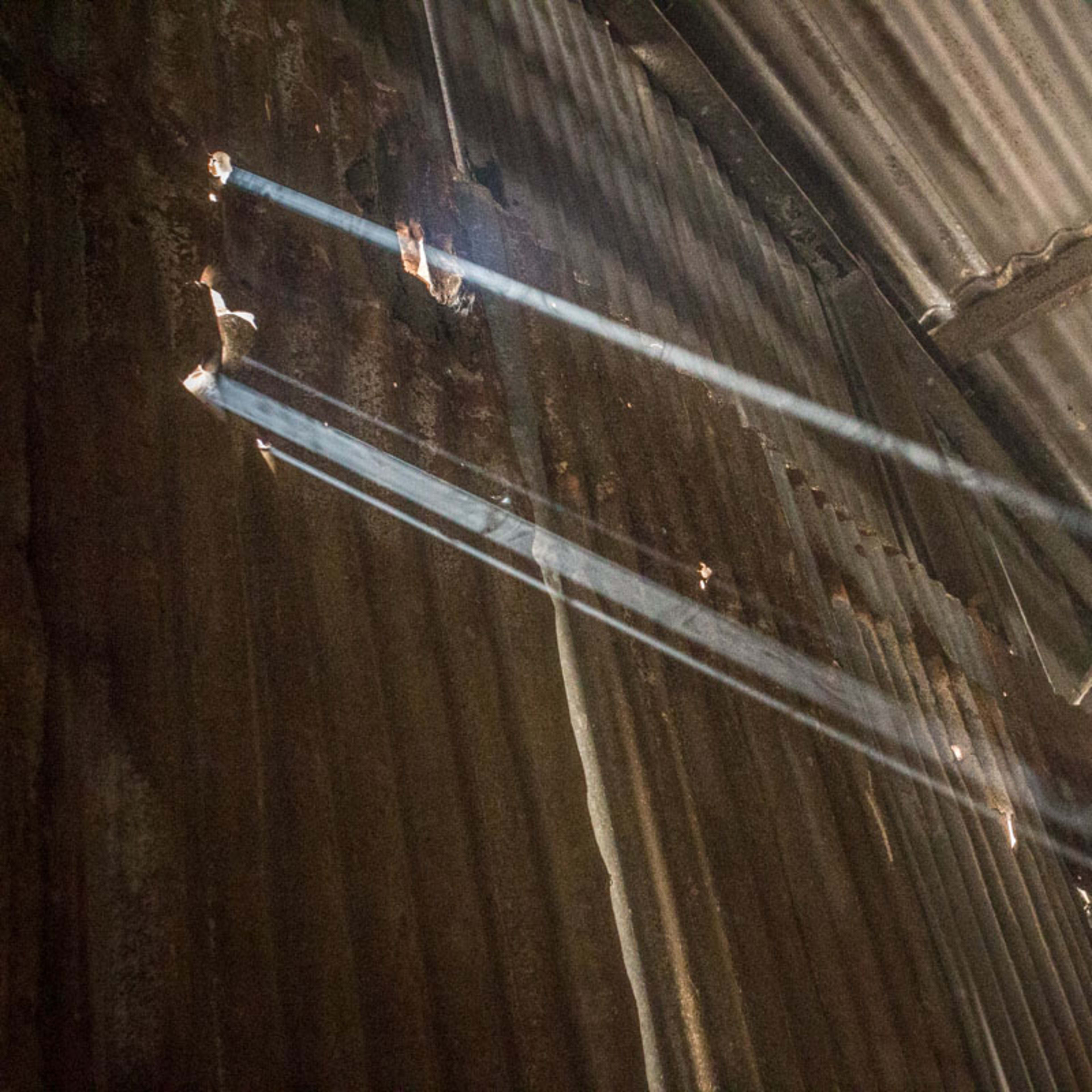 Sunlight filters through gaps in a corrugated metal roof, creating beams of light against the rusty walls of an interior space.