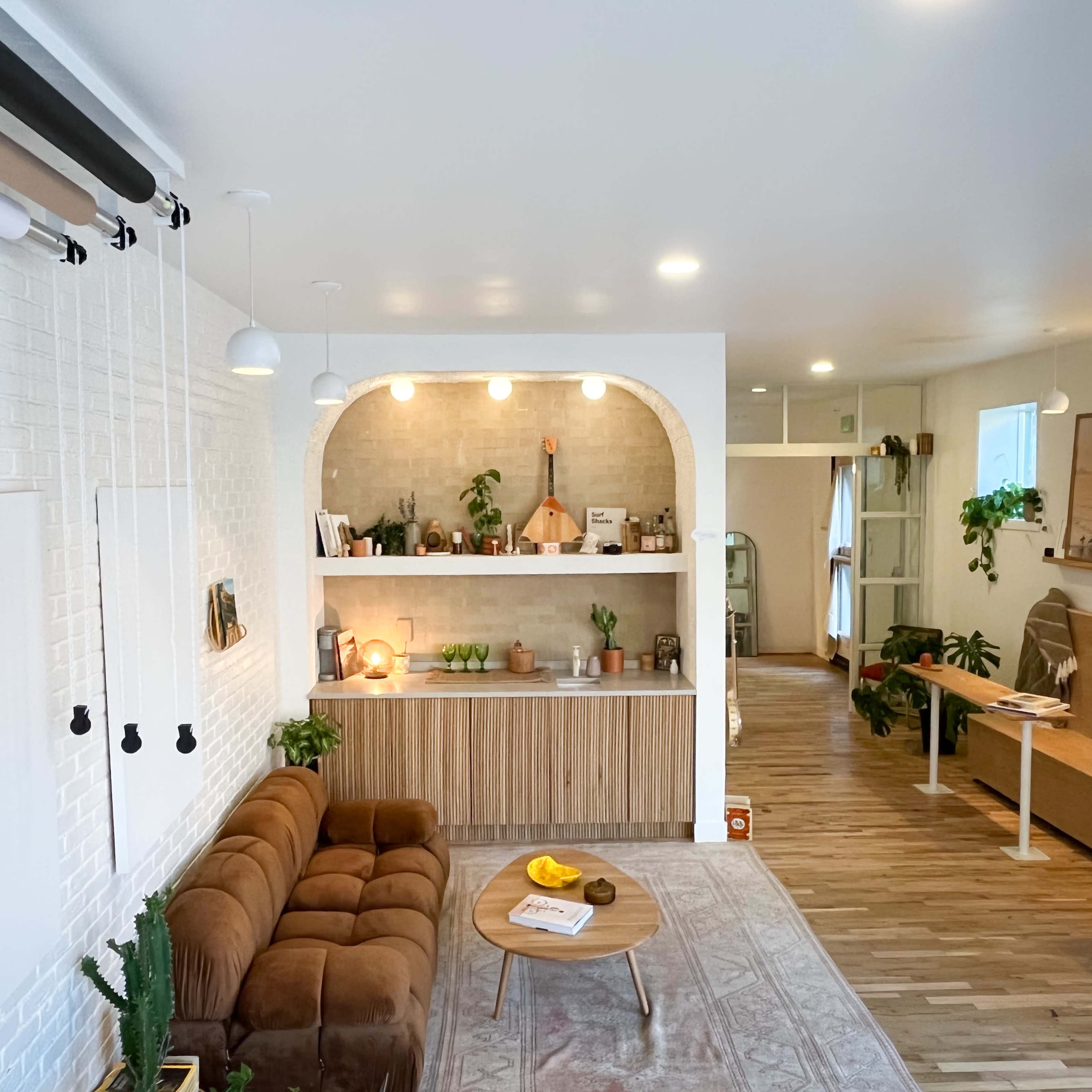 A modern, open-concept living space with a brown sofa, a central wooden table, and a kitchen area featuring plants and various decorative items.