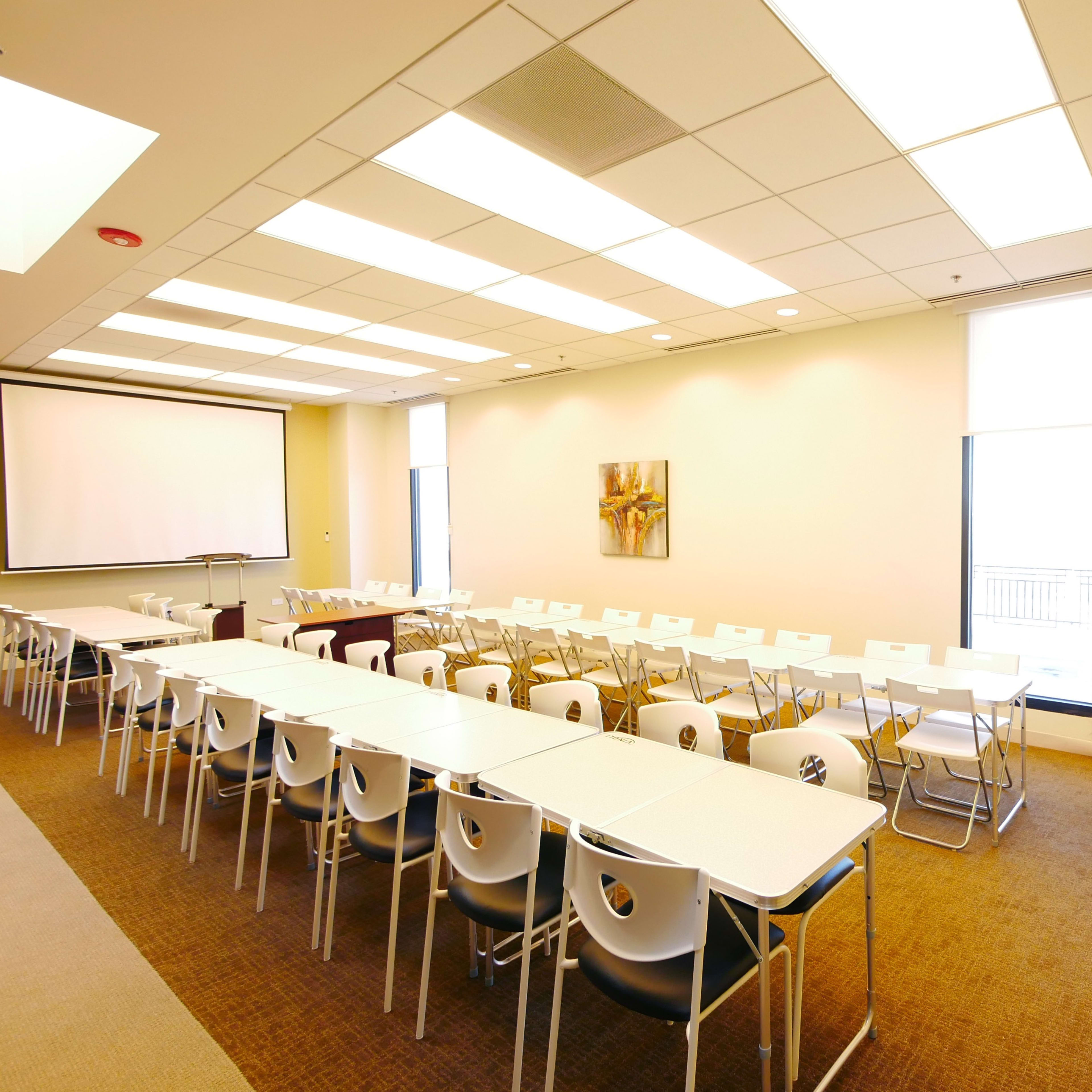A well-lit meeting room with multiple tables arranged in a U-shape and rows of white chairs along the walls.