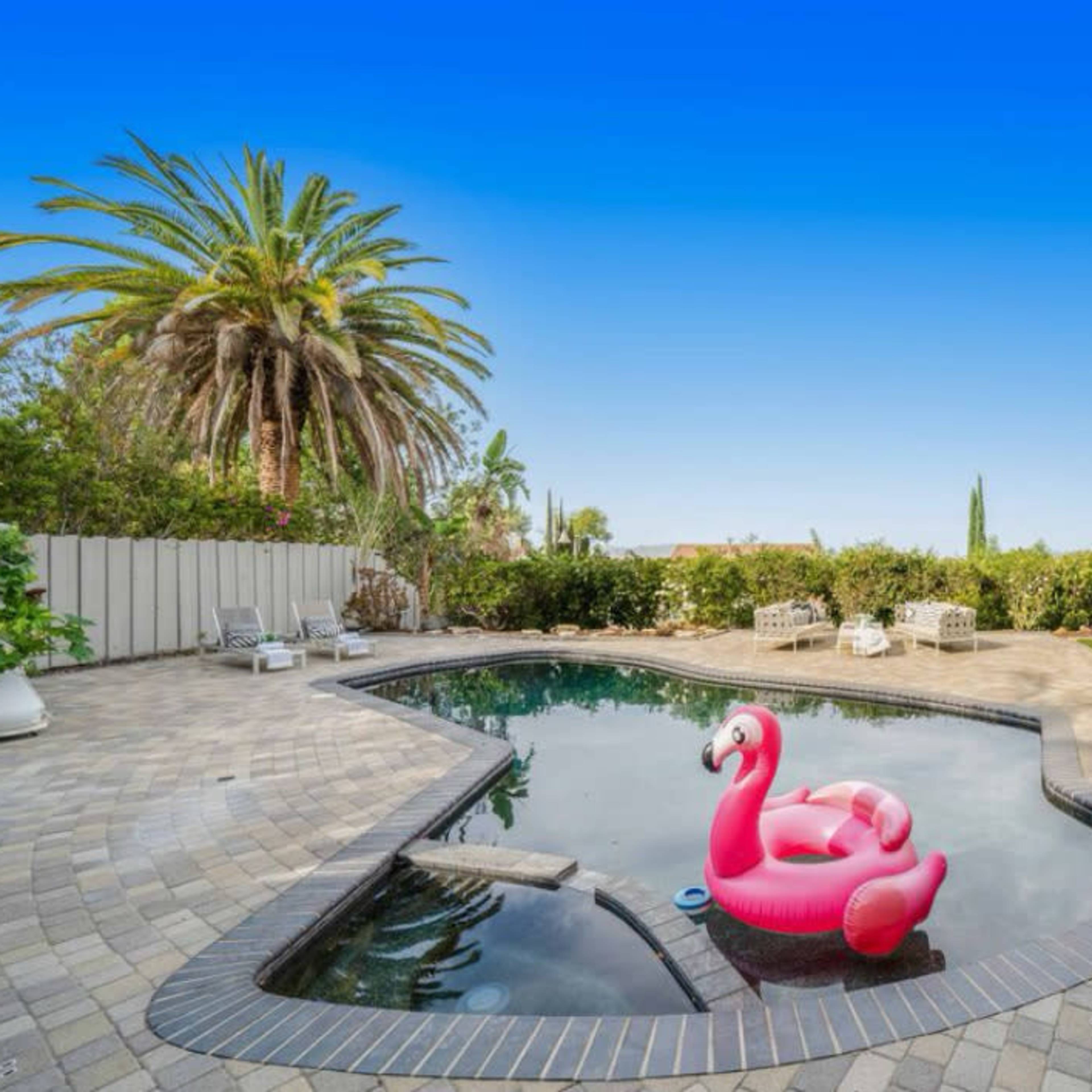 A pink flamingo float sits in a swimming pool surrounded by palm trees and patio furniture.