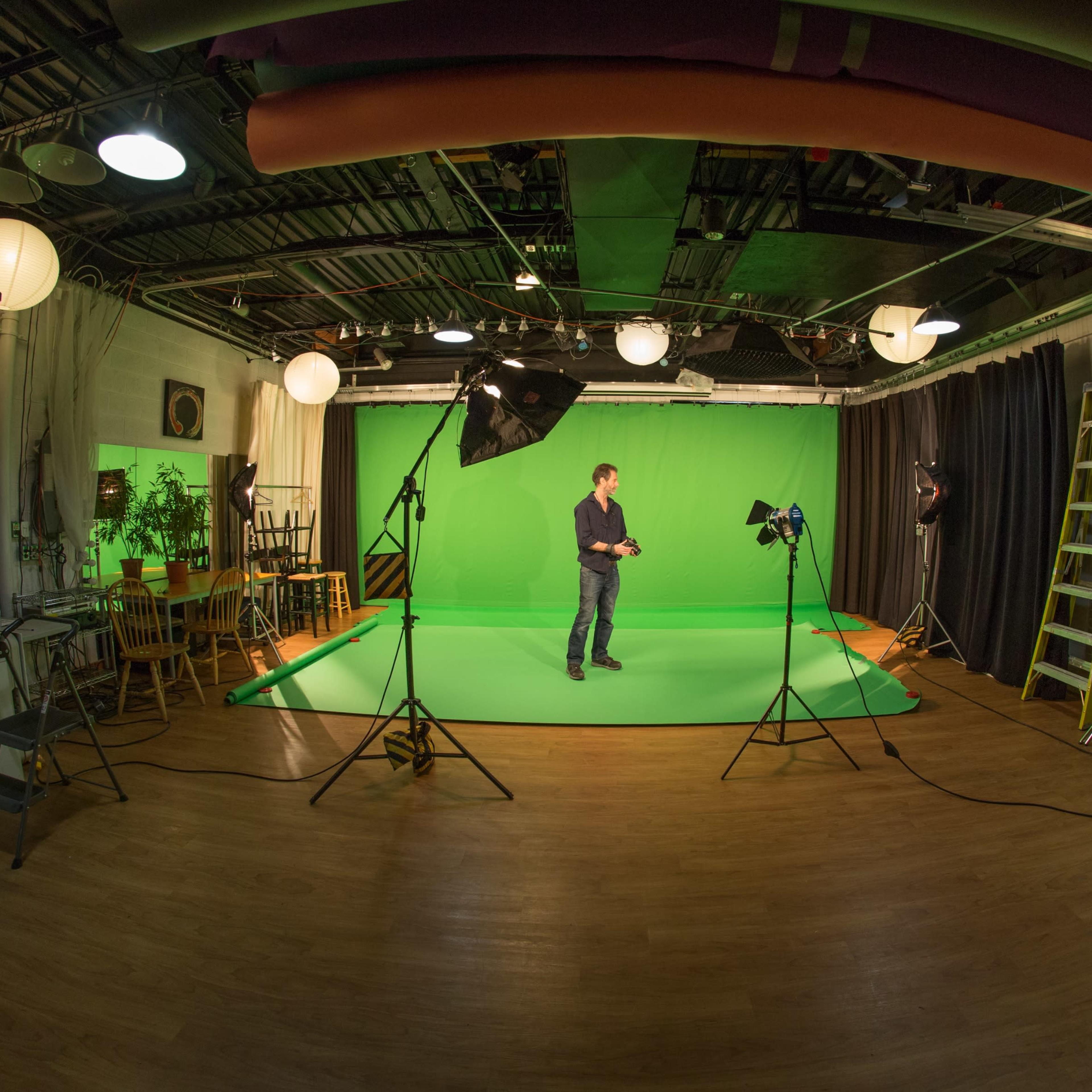 A man stands in a studio with green screens, studio lights, and various equipment arranged around him.