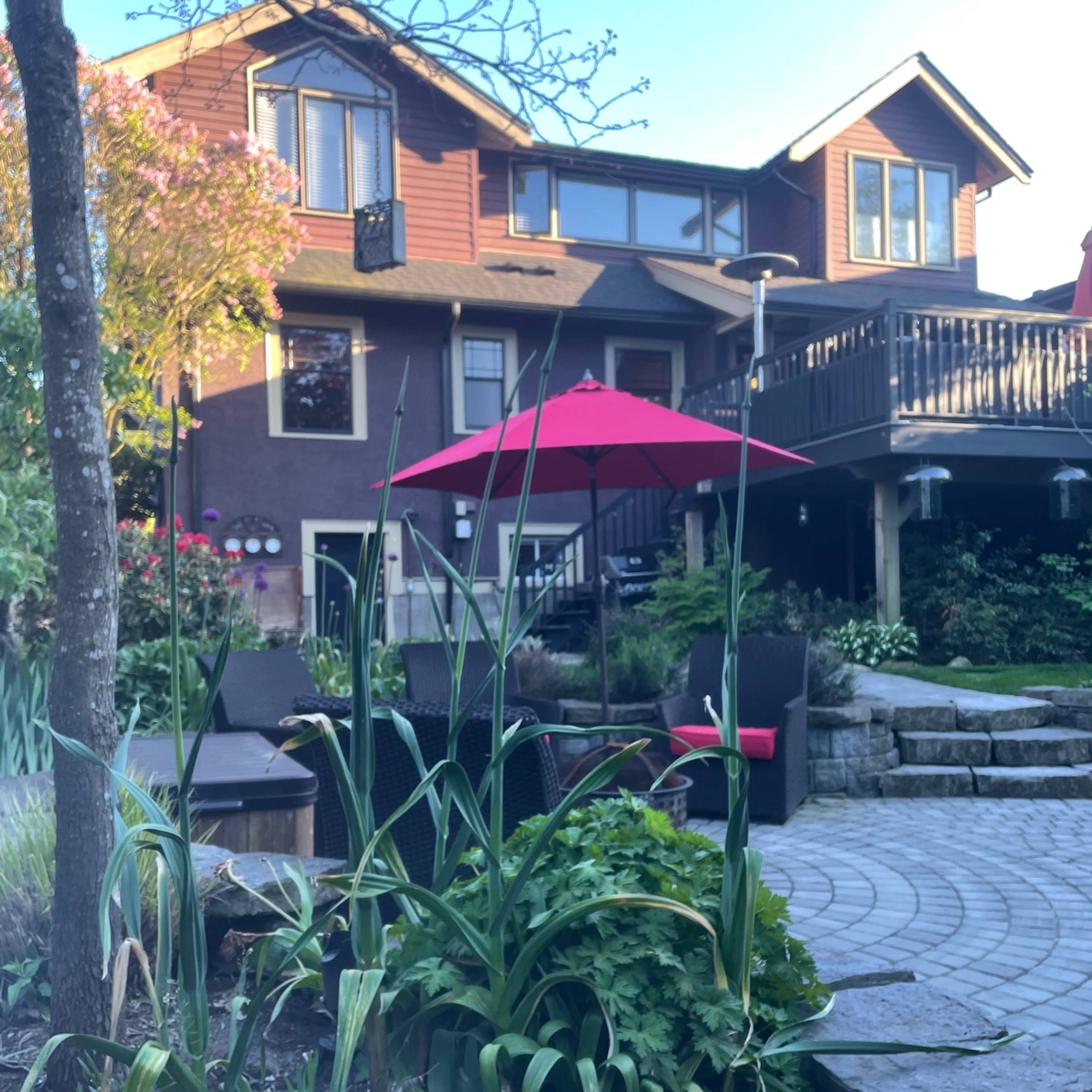 A patio area with outdoor furniture and a red umbrella is situated in front of a two-story brown house, surrounded by landscaped plants and stone pathways.