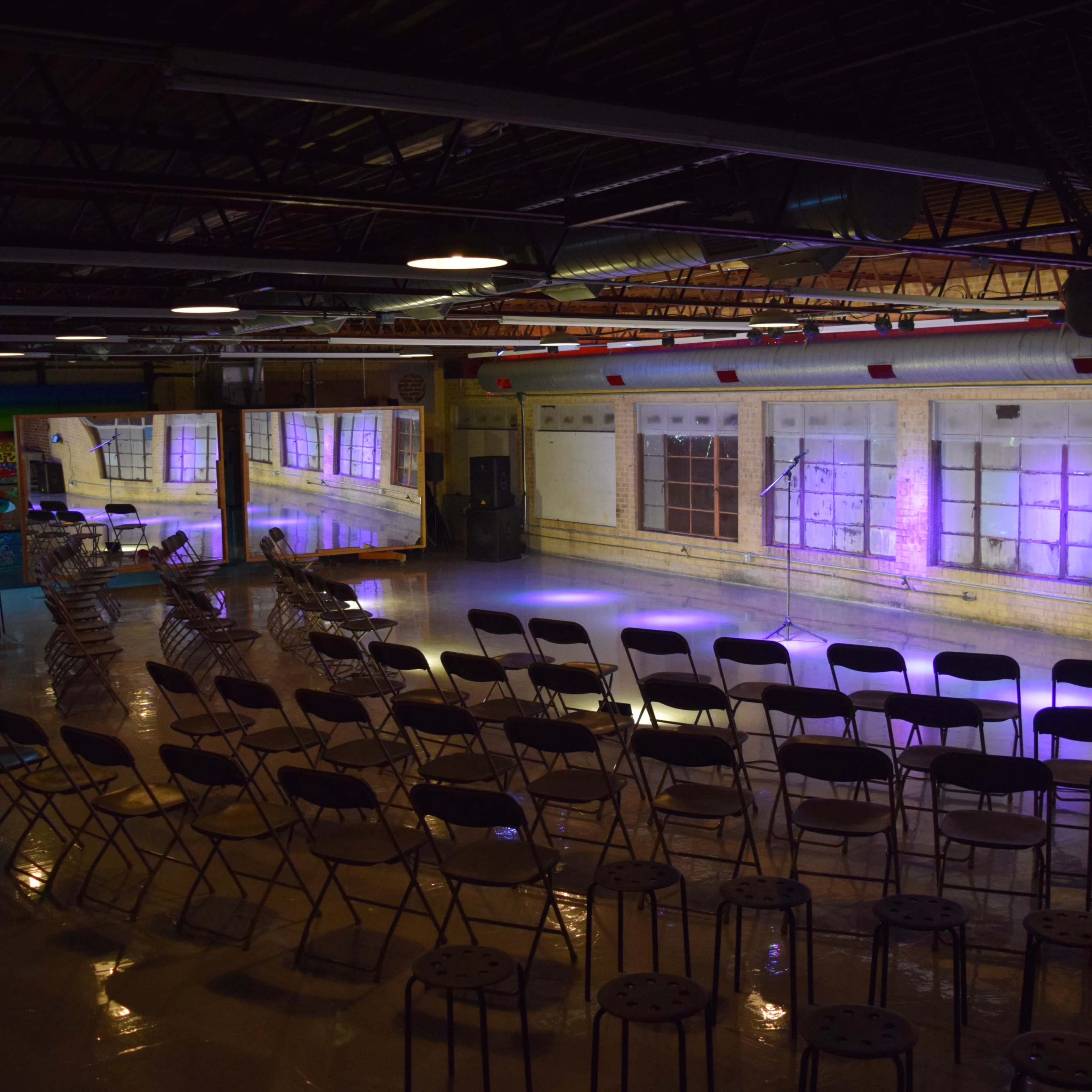 A spacious indoor venue set up with rows of chairs facing a stage, illuminated by soft purple lighting against a backdrop of large windows.