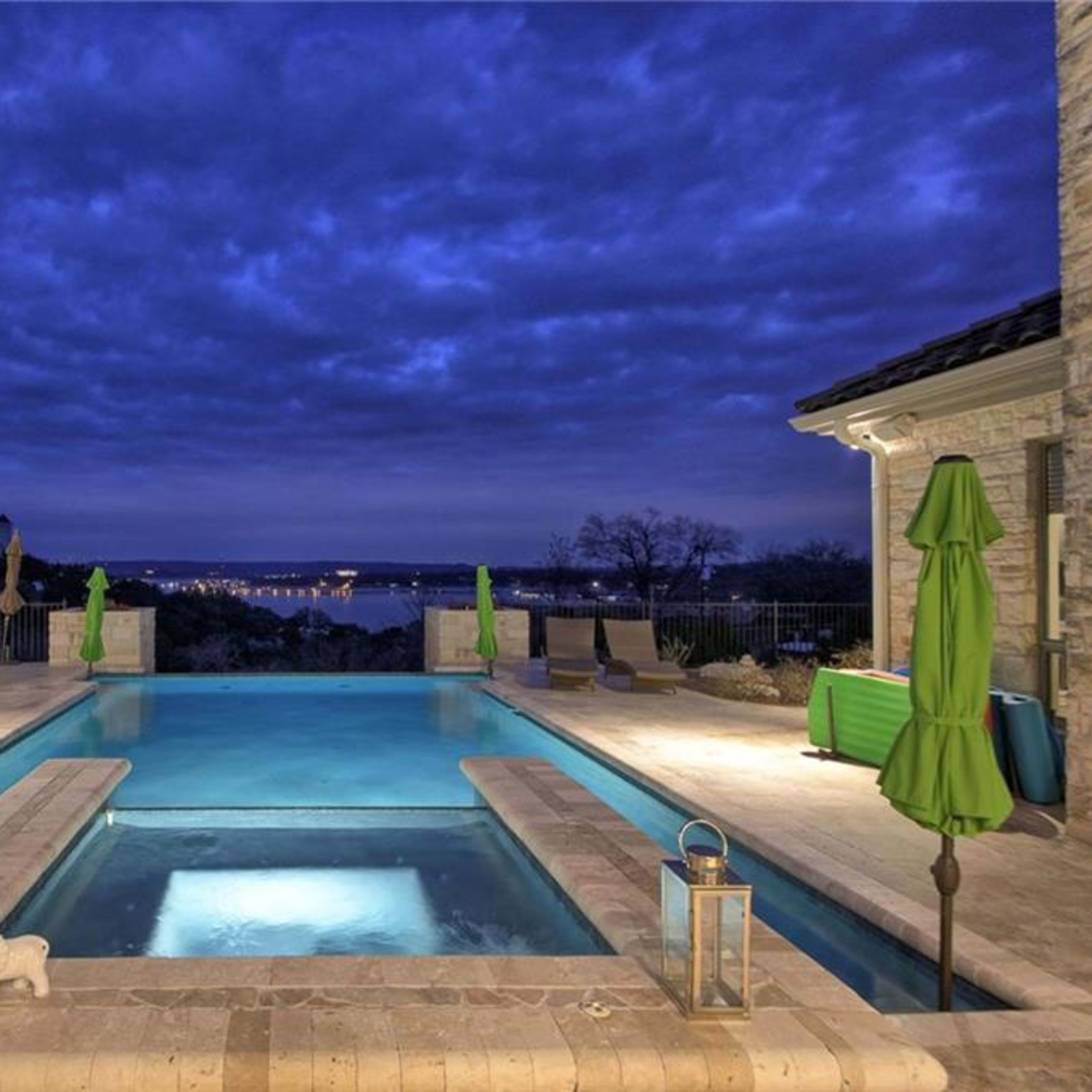 A well-lit pool area features green umbrellas and overlooks a distant skyline under a cloudy twilight sky.