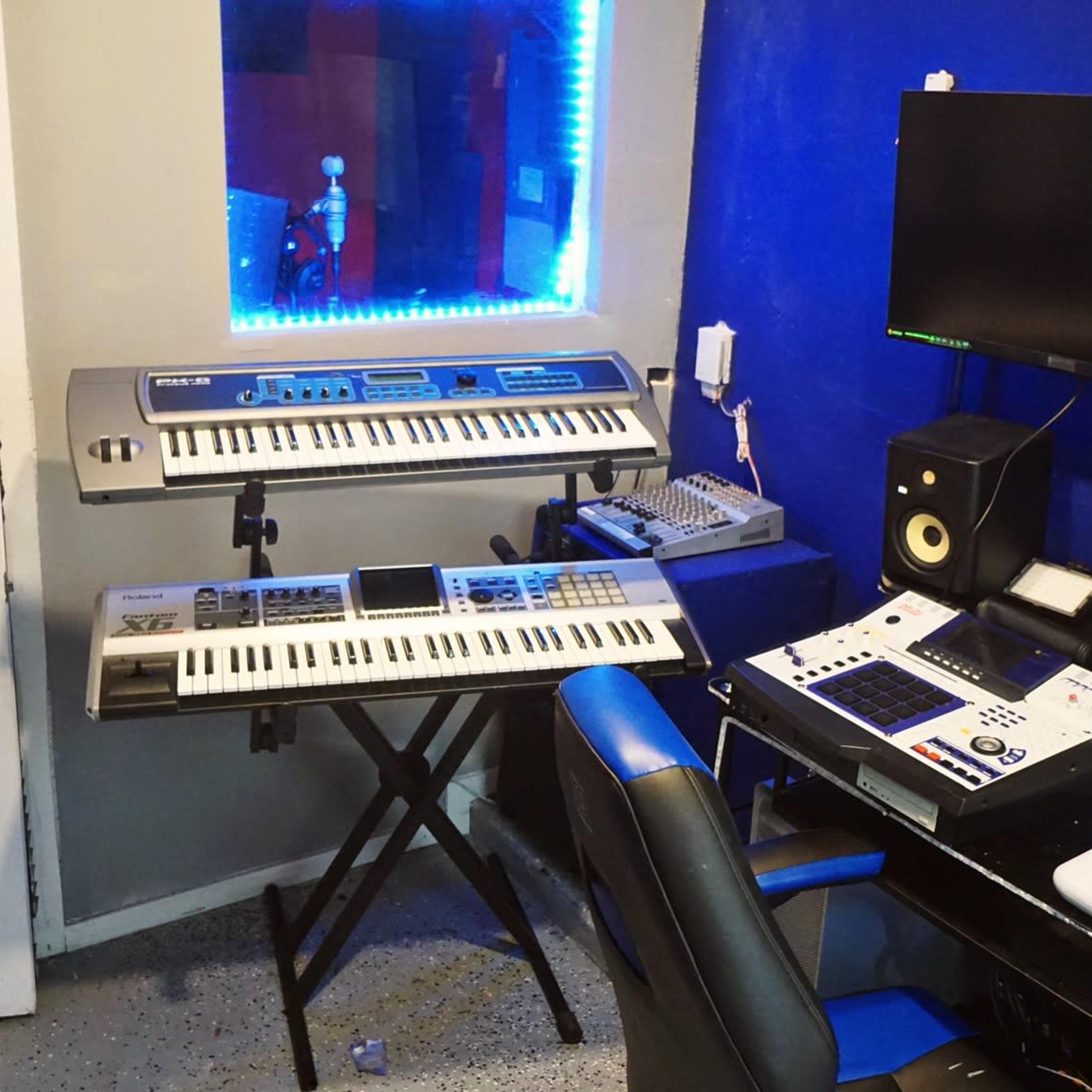 A music production setup in a room, featuring two keyboards, a computer station with multiple monitors, and a door.