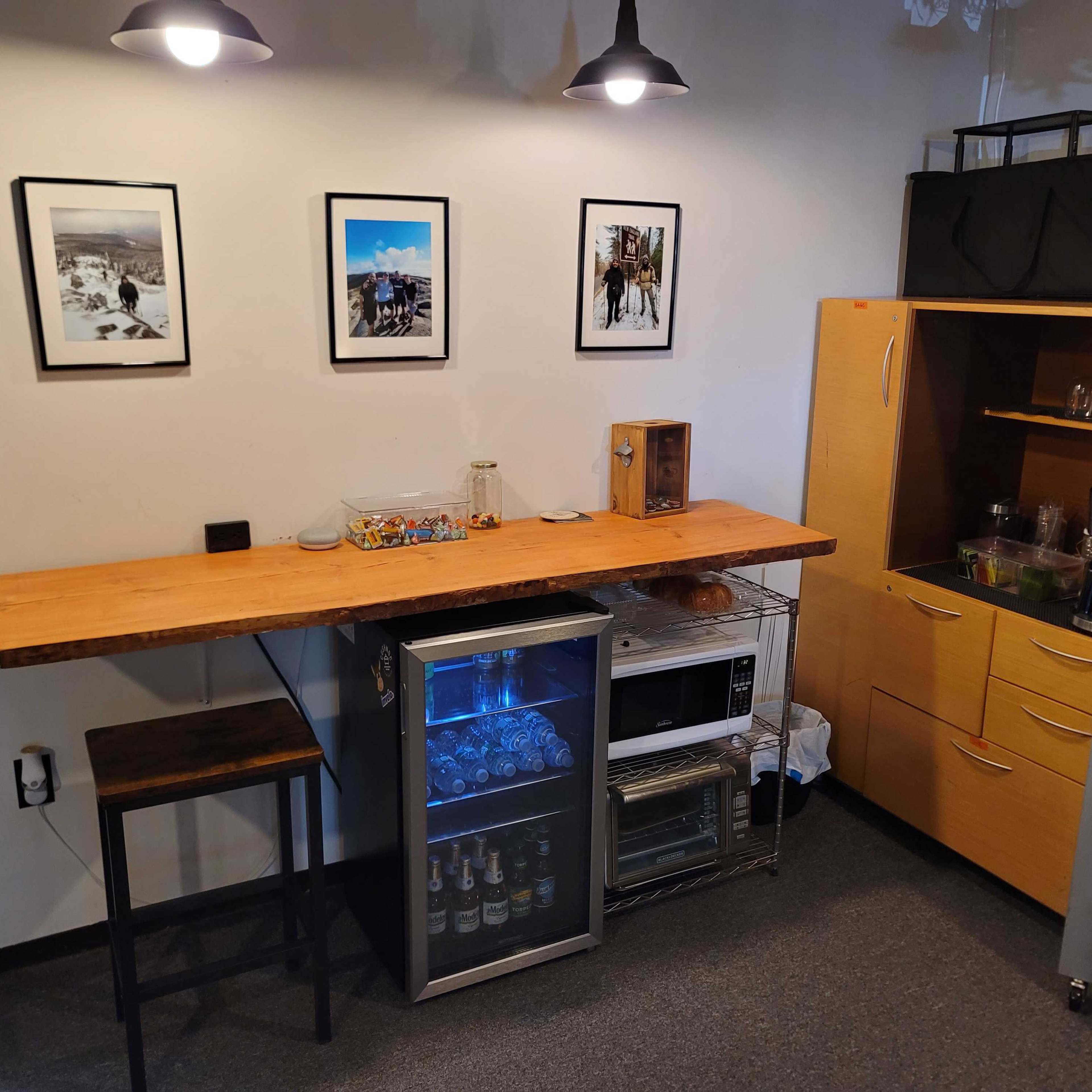 A small kitchen space with a wooden bar counter, framed pictures on the wall, and a glass-front refrigerator beneath the counter.