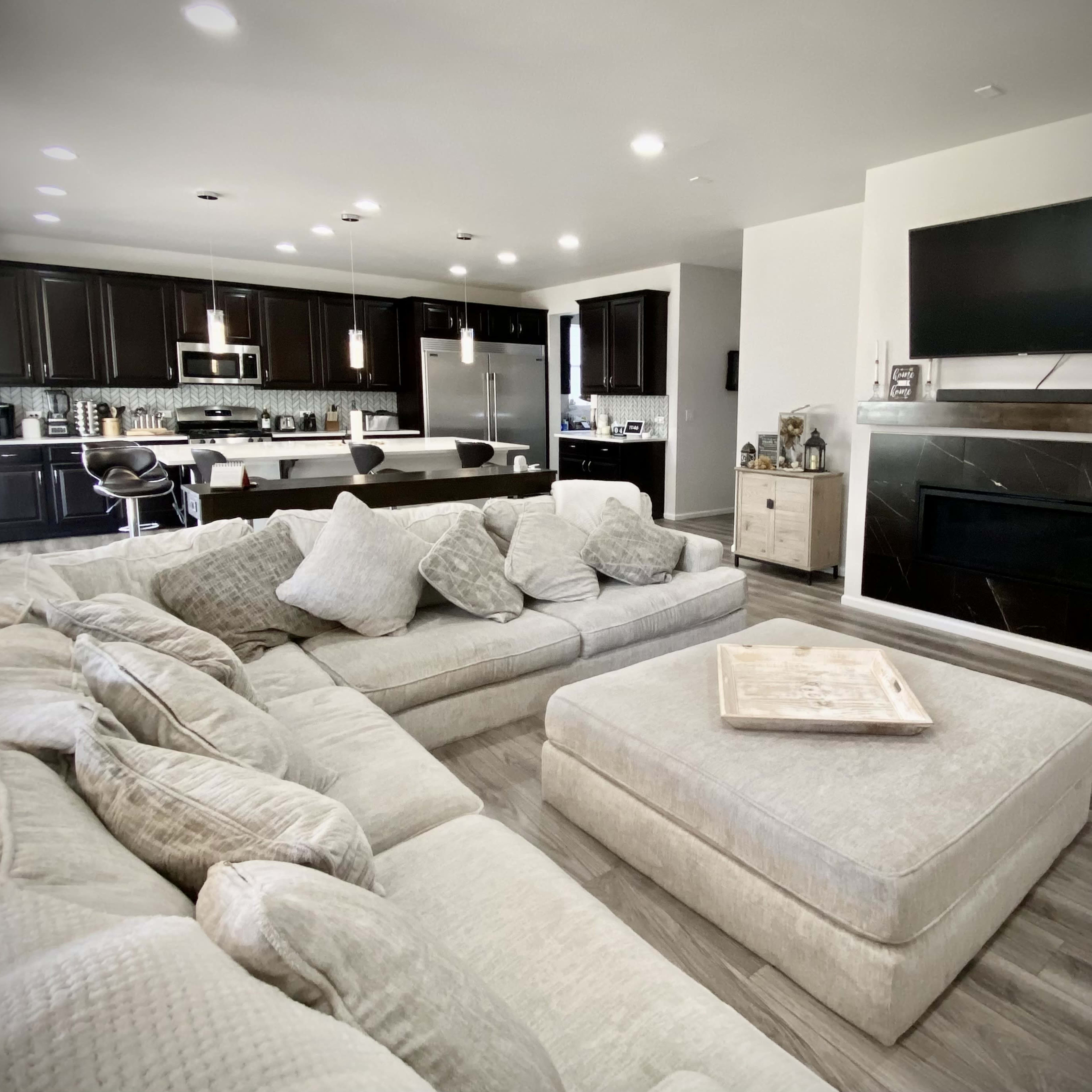 A modern living room featuring a large sectional sofa, an open kitchen with dark cabinetry, and a flat-screen TV mounted above a fireplace.