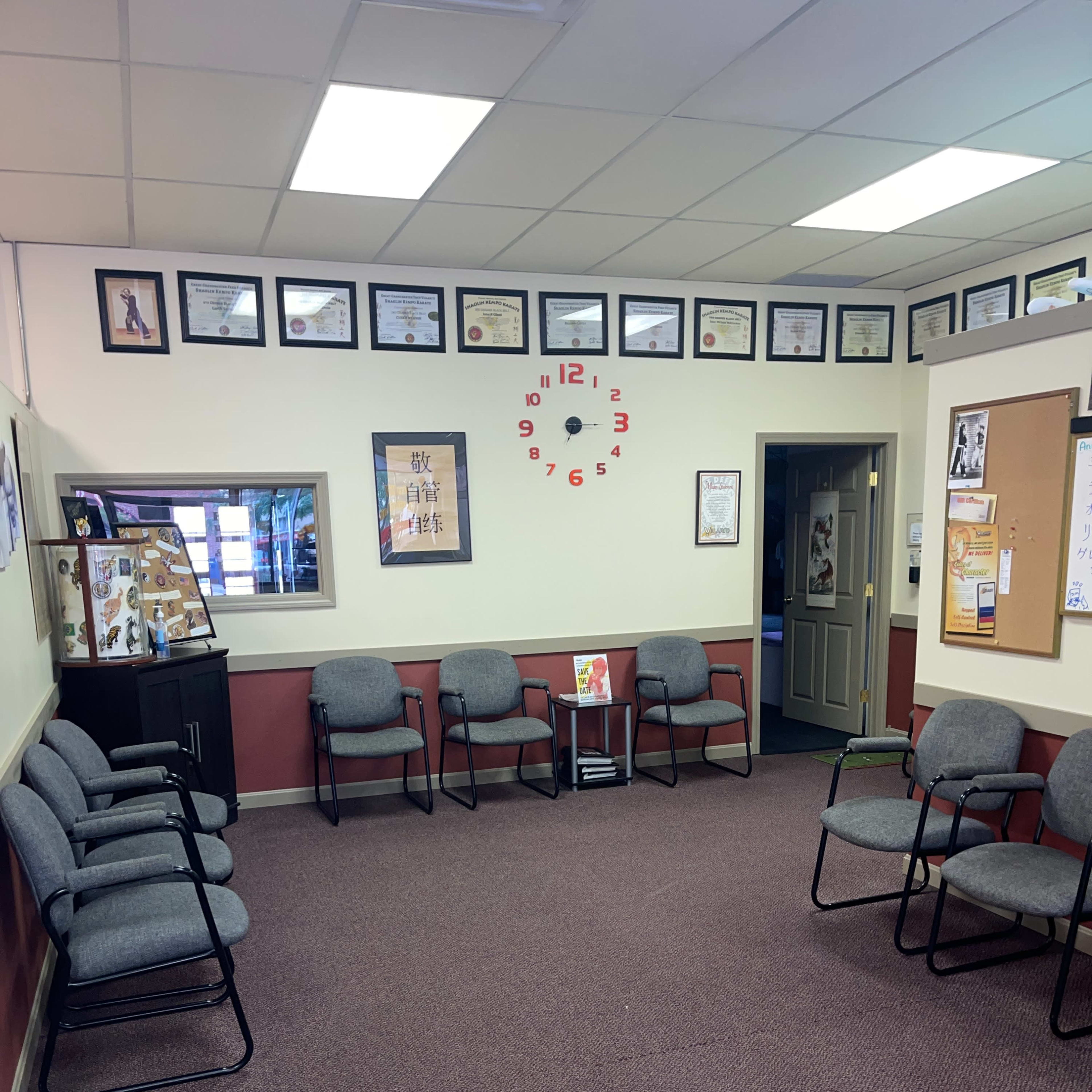 A waiting area with several gray chairs arranged around a central space, featuring framed certificates and a clock on the wall.