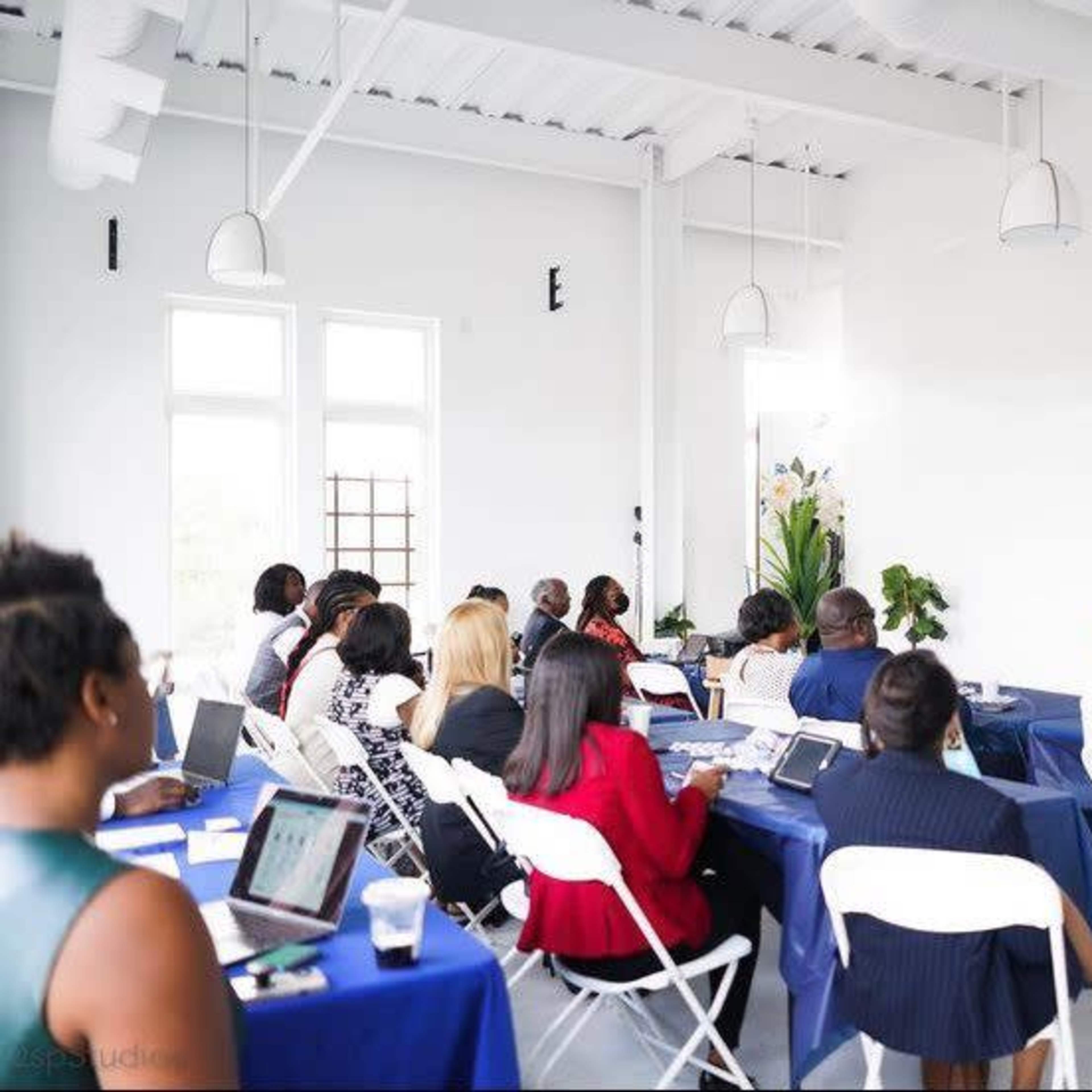 A speaker presents to an audience seated at tables in a modern conference room.