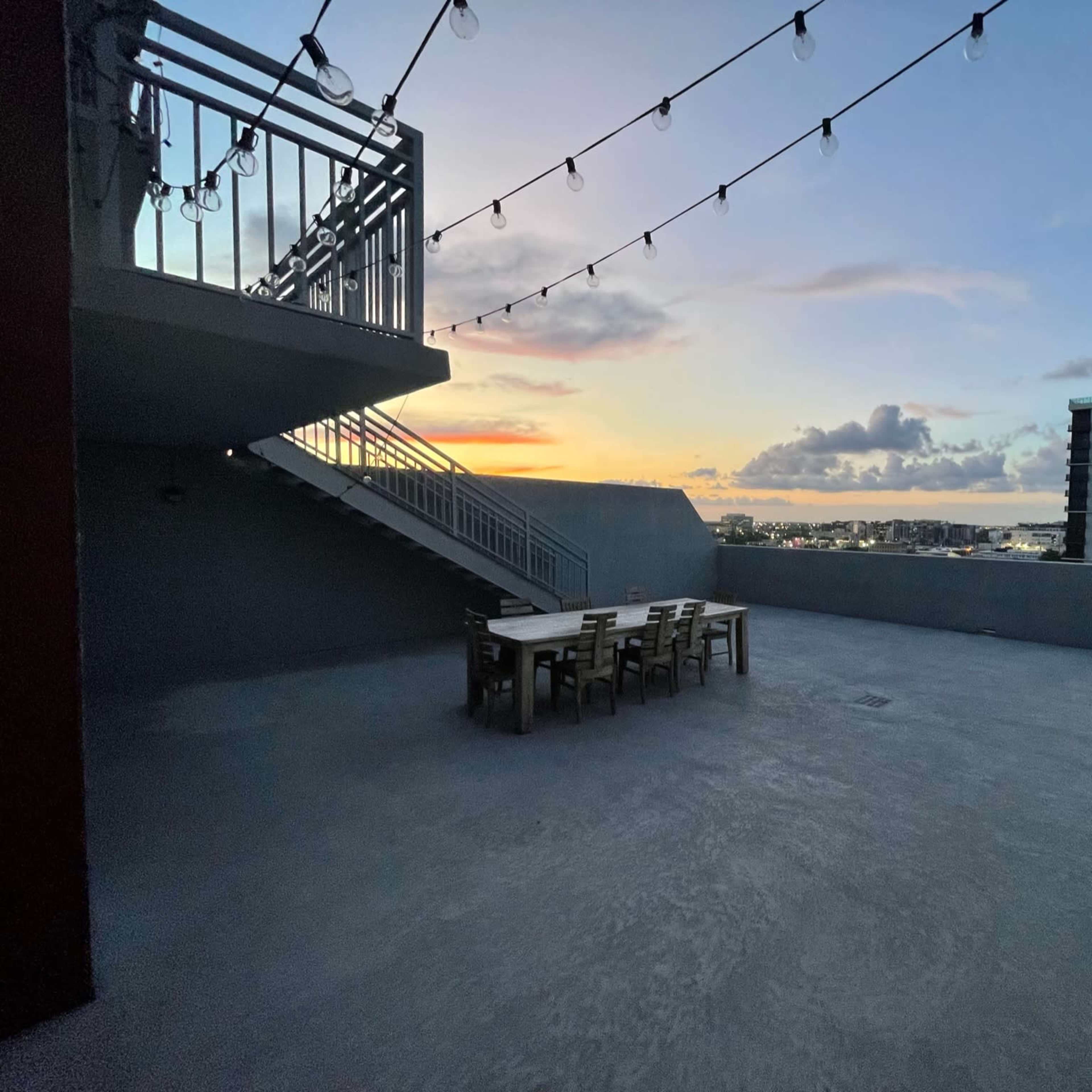 A rooftop terrace with a dining table and chairs, illuminated by string lights, overlooking a city skyline at sunset.