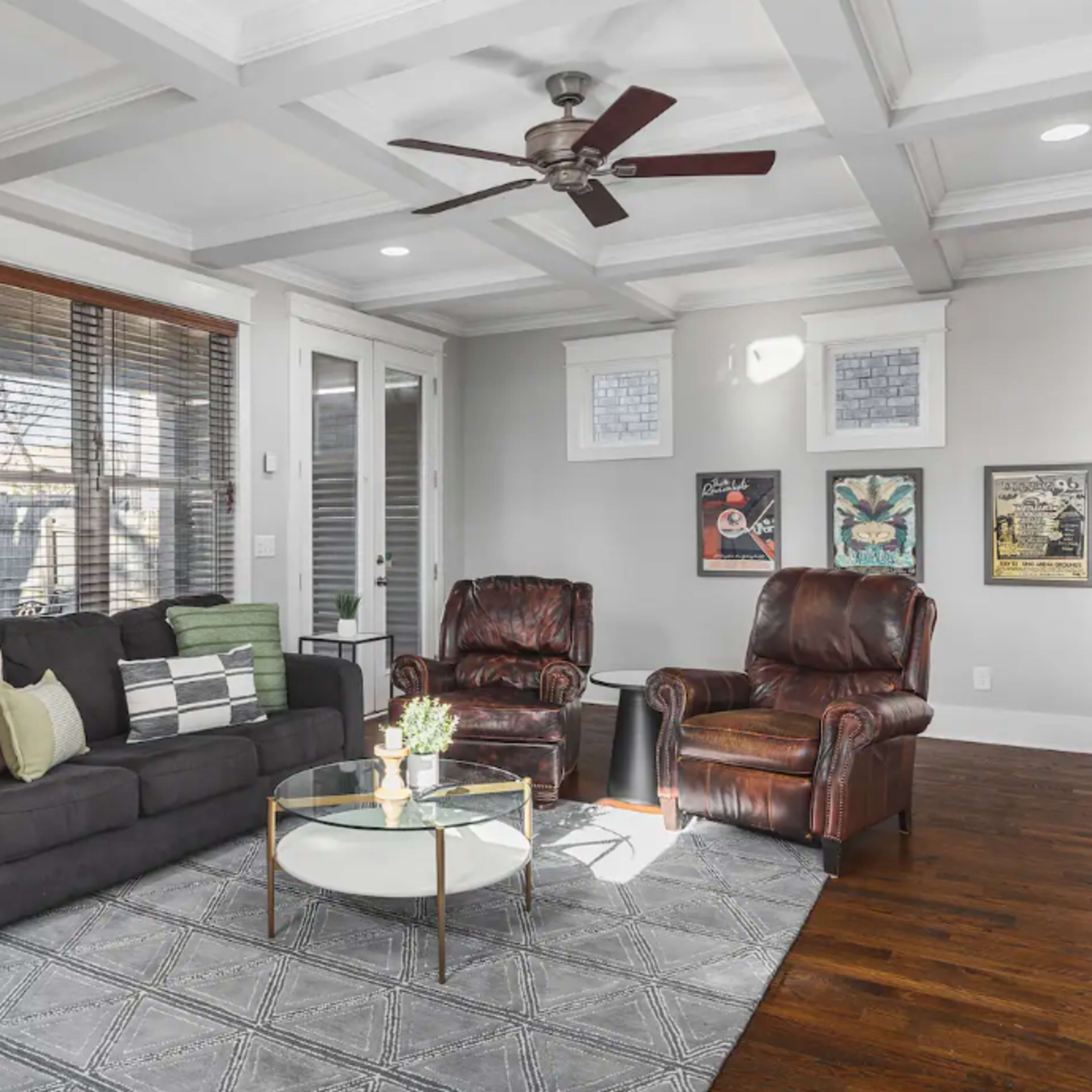 The living room features a black sofa, two brown recliners, a round coffee table, and large windows covered with wooden blinds.