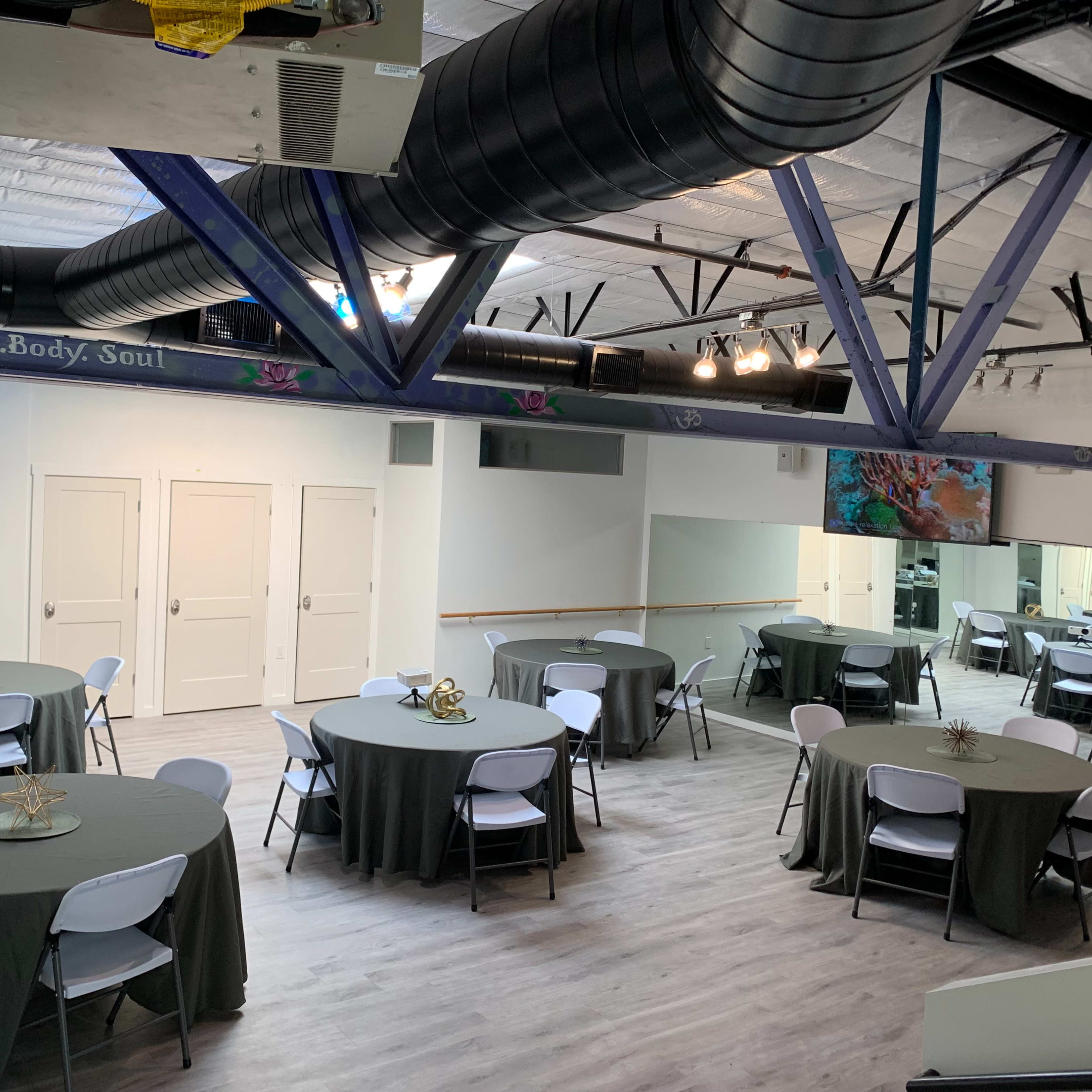 A spacious room with several round tables covered in gray tablecloths, set up for an event, and featuring large mirrors and a mounted screen on the wall.