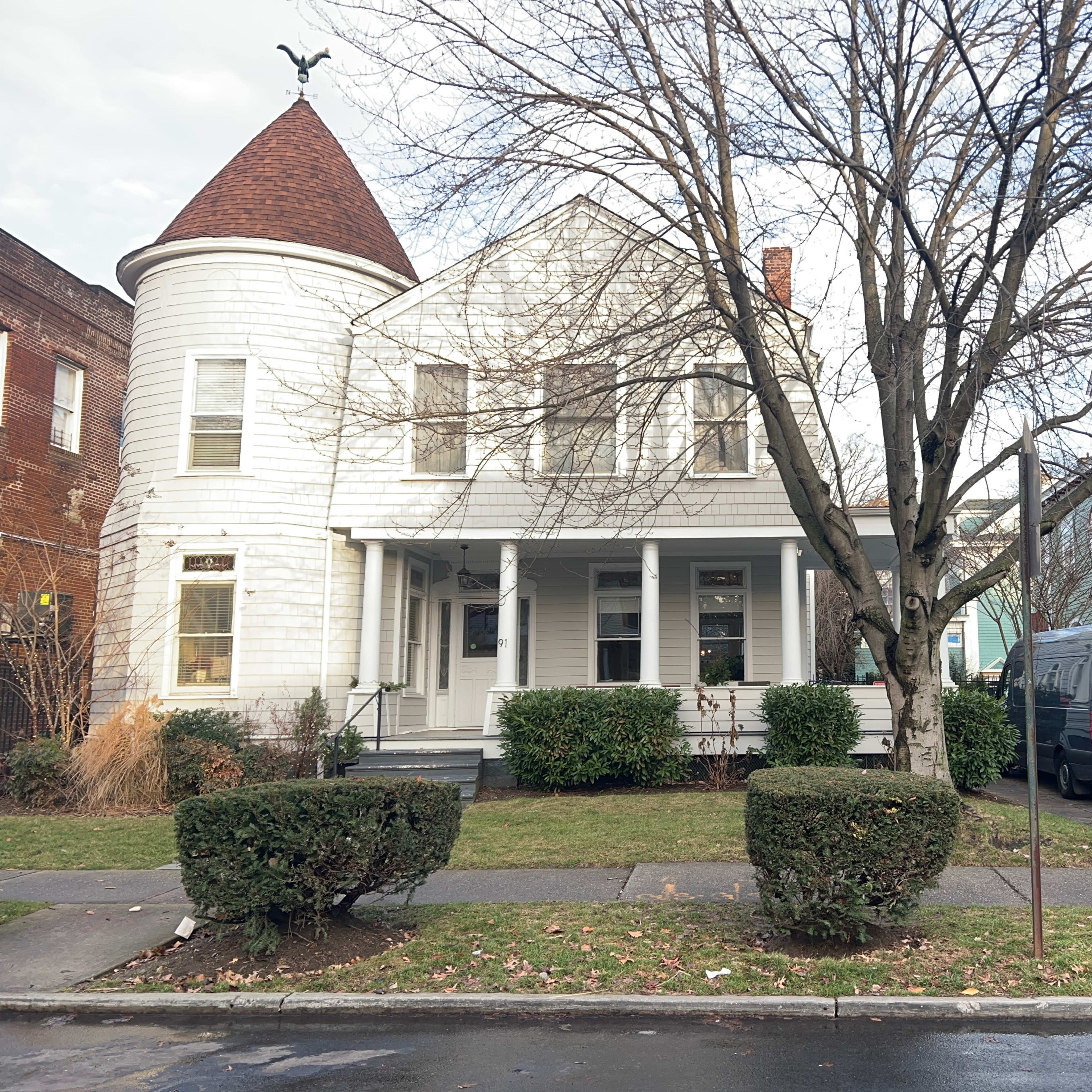 A white two-story house with a pointed roof and a large front porch is situated next to a green house, with a black vehicle parked in the driveway.