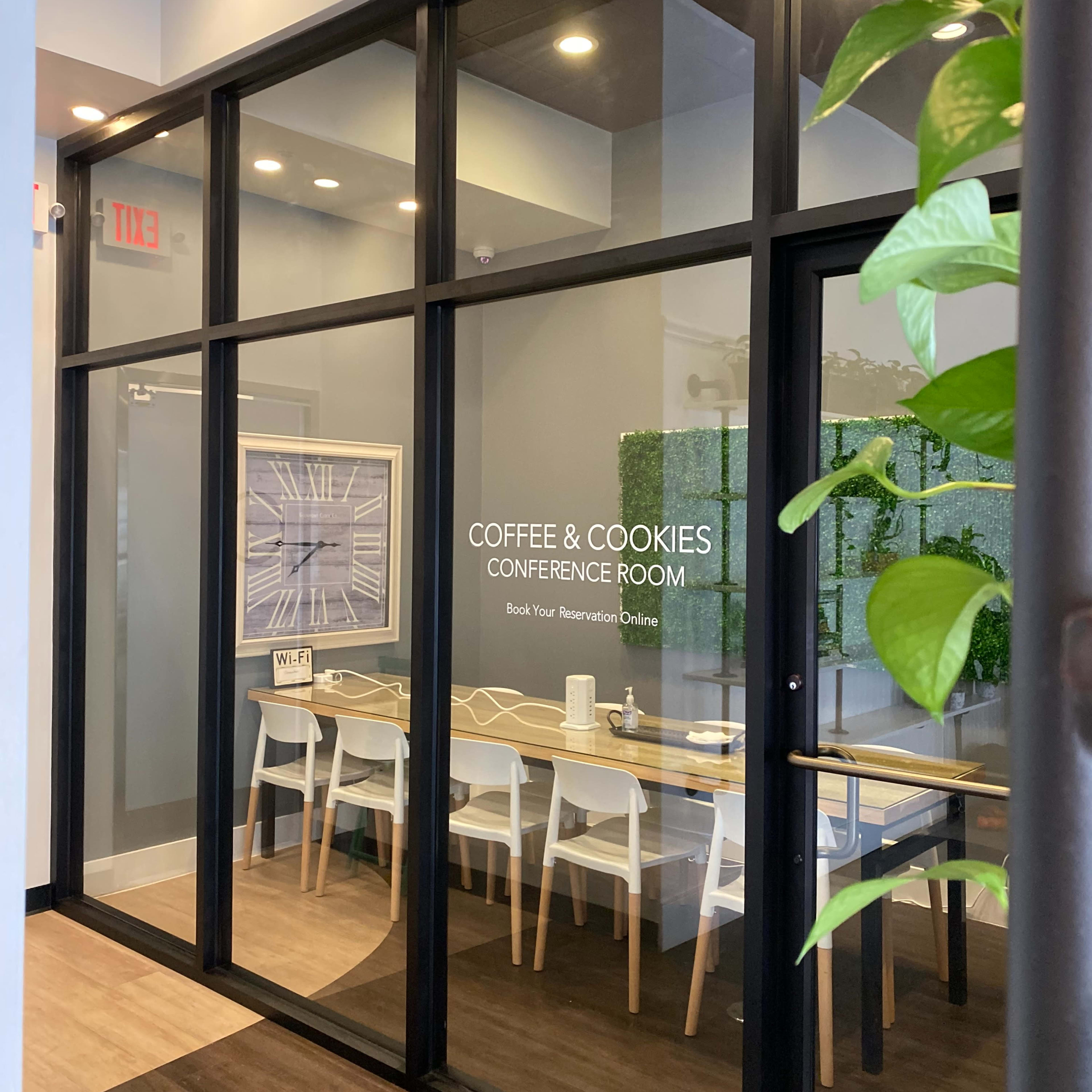A glass-walled conference room titled "Coffee & Cookies" features a long table surrounded by white chairs and greenery decor.