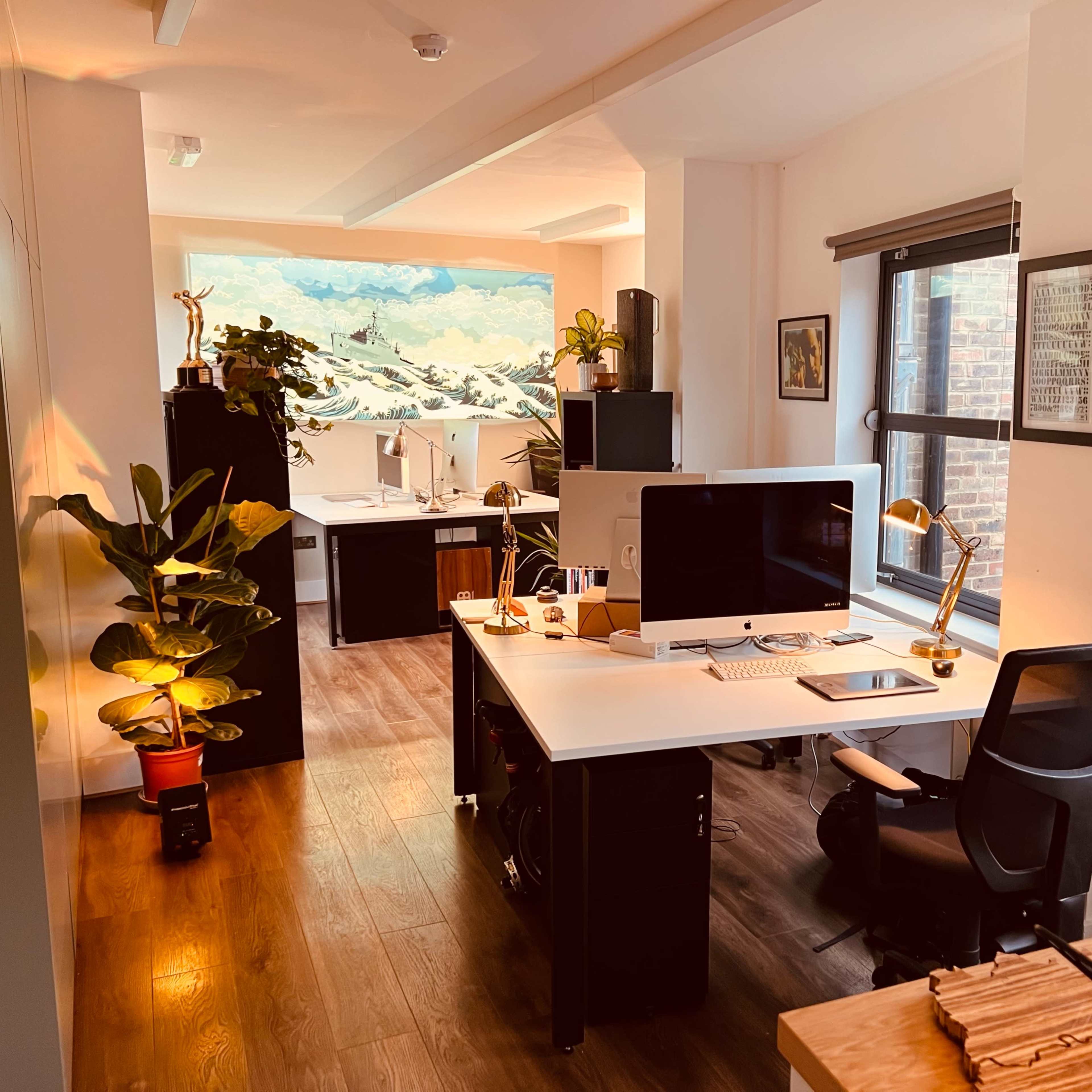 A modern office space with two workstations, featuring desks with computers, plants, and artwork on the walls.