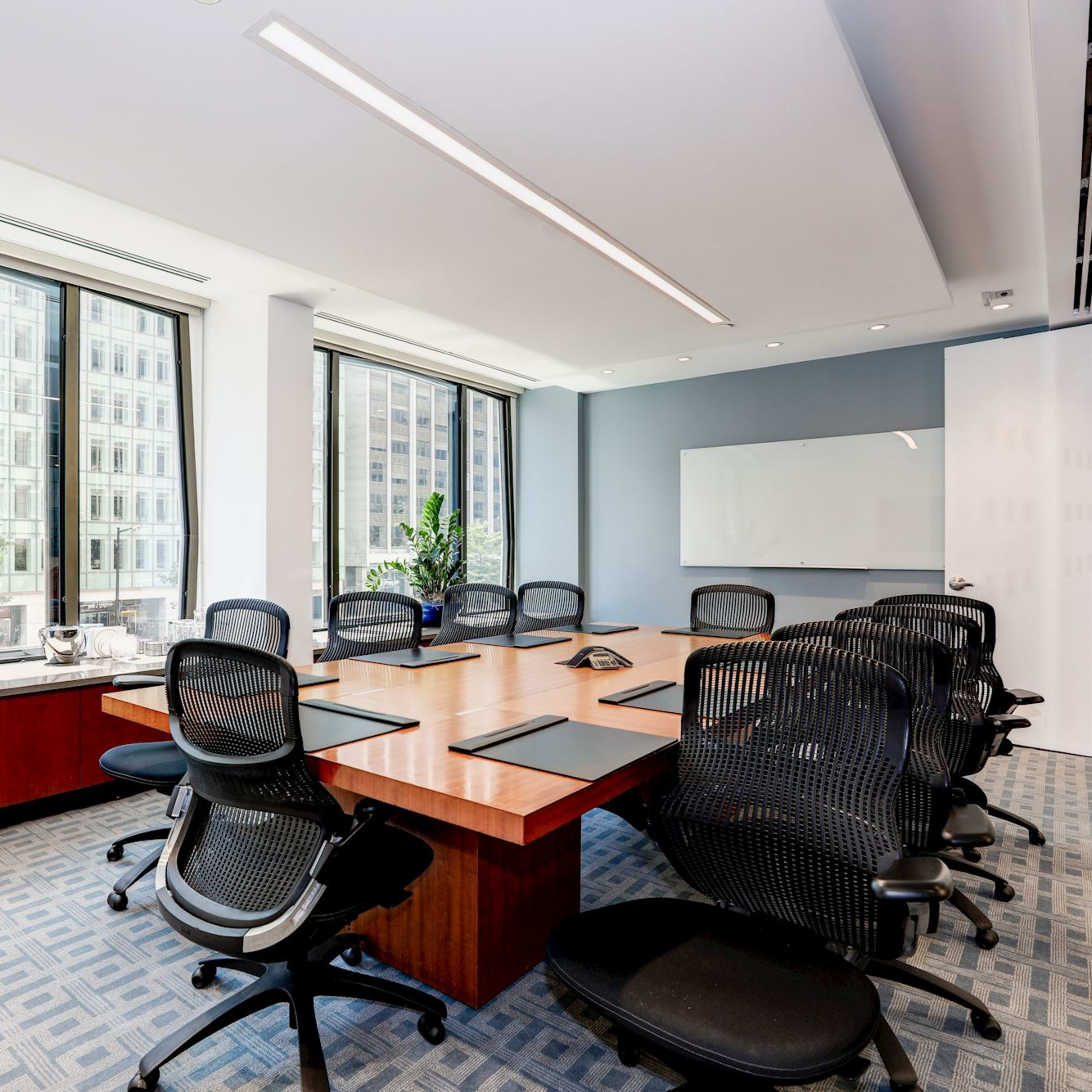 A conference room with a large wooden table surrounded by black ergonomic chairs, featuring large windows that provide a view of a cityscape and potted plants in the corners.