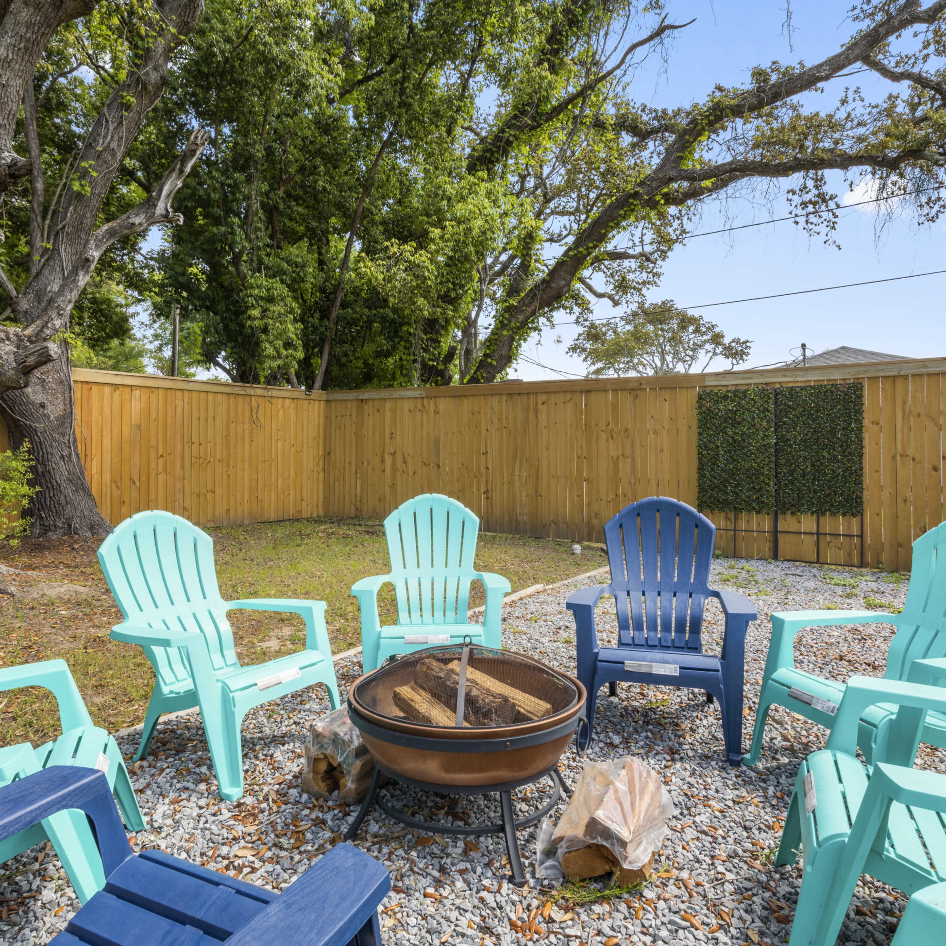A fire pit surrounded by eight colorful adirondack chairs is set in a grass area enclosed by a wooden fence.