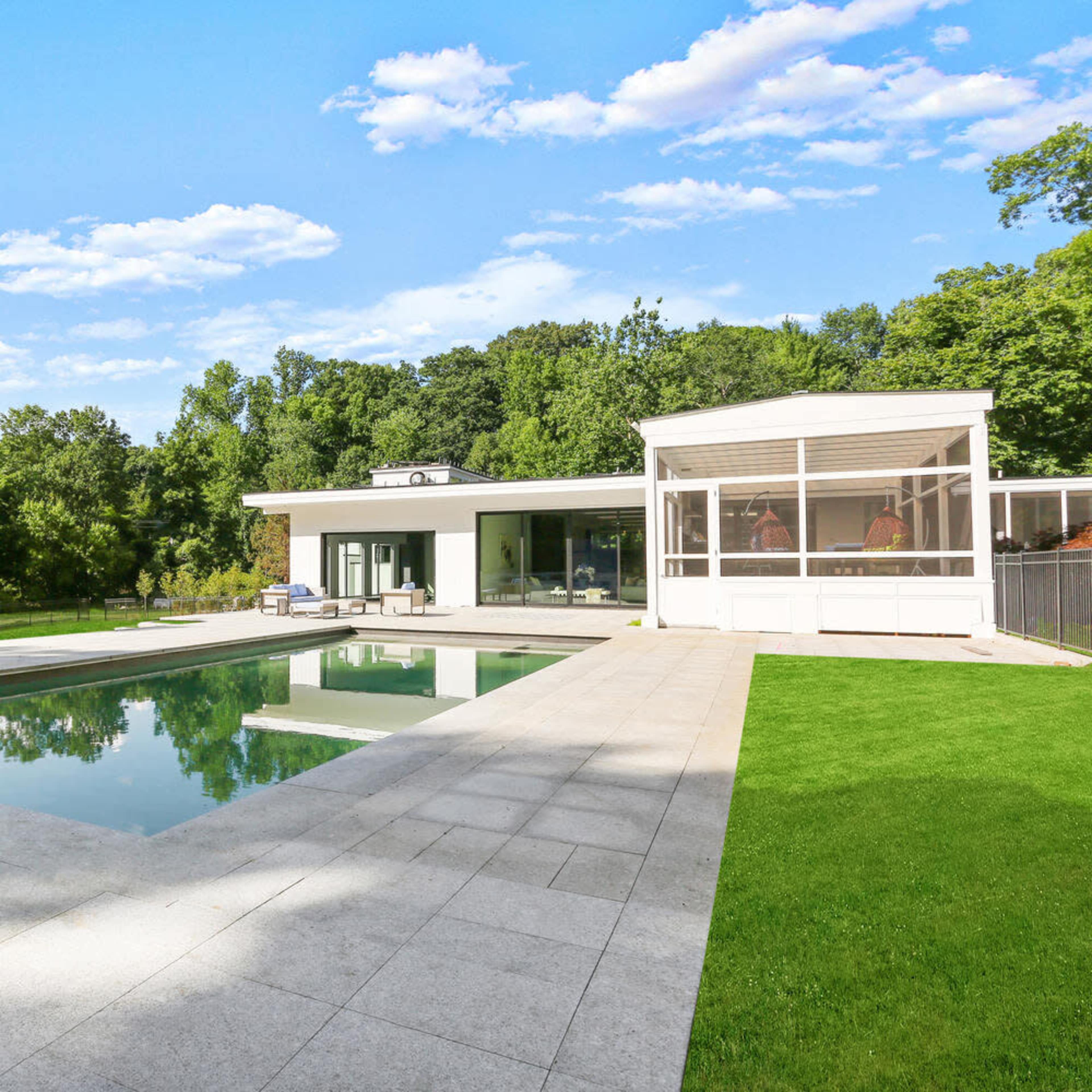 A modern house with large glass windows and a swimming pool surrounded by a stone patio and green lawn, set against a backdrop of trees.