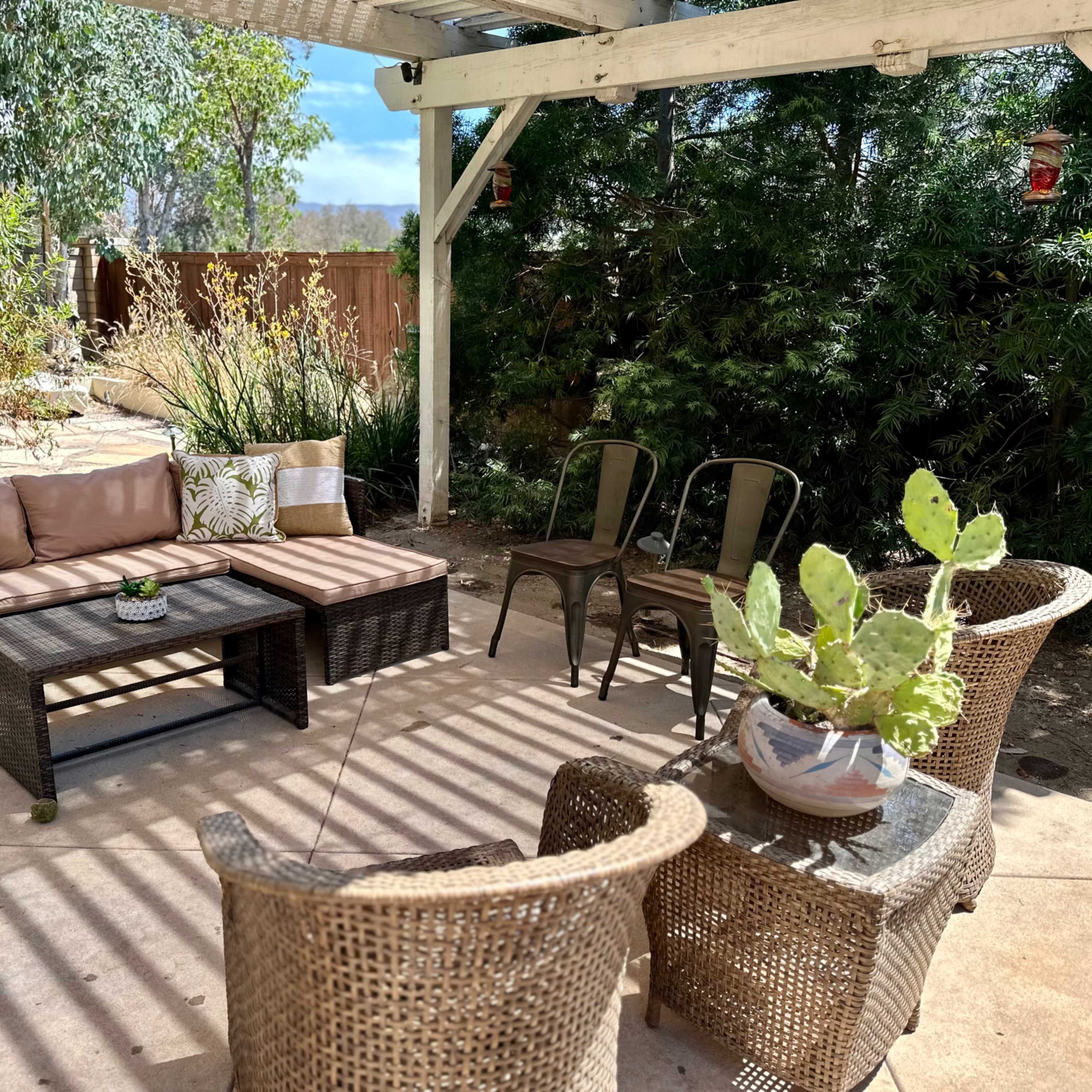 A patio area features a sectional sofa, two chairs, a table, and potted plants underneath a shaded structure.