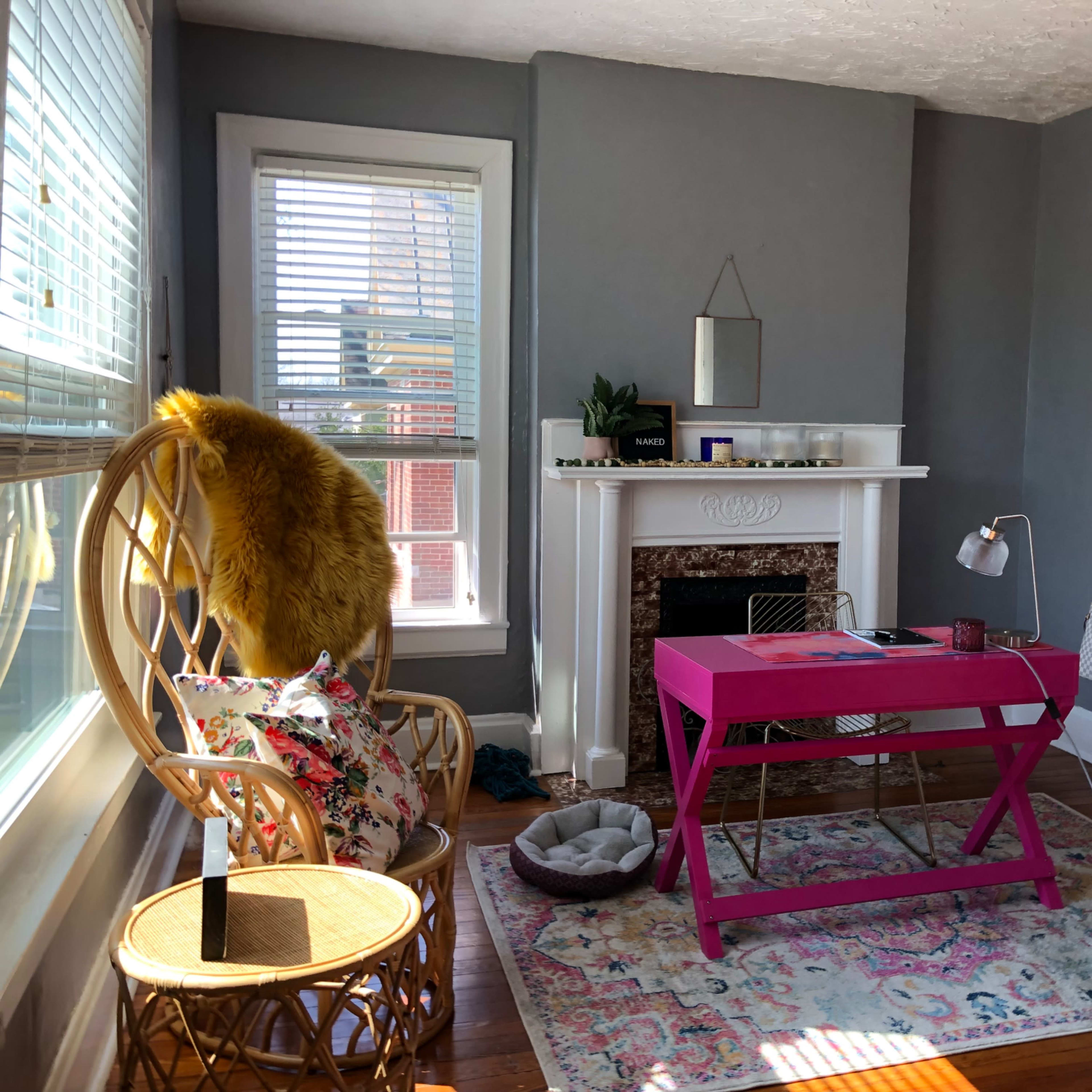 A brightly lit room with a pink desk, a rattan chair, and a decorative rug in front of a fireplace.