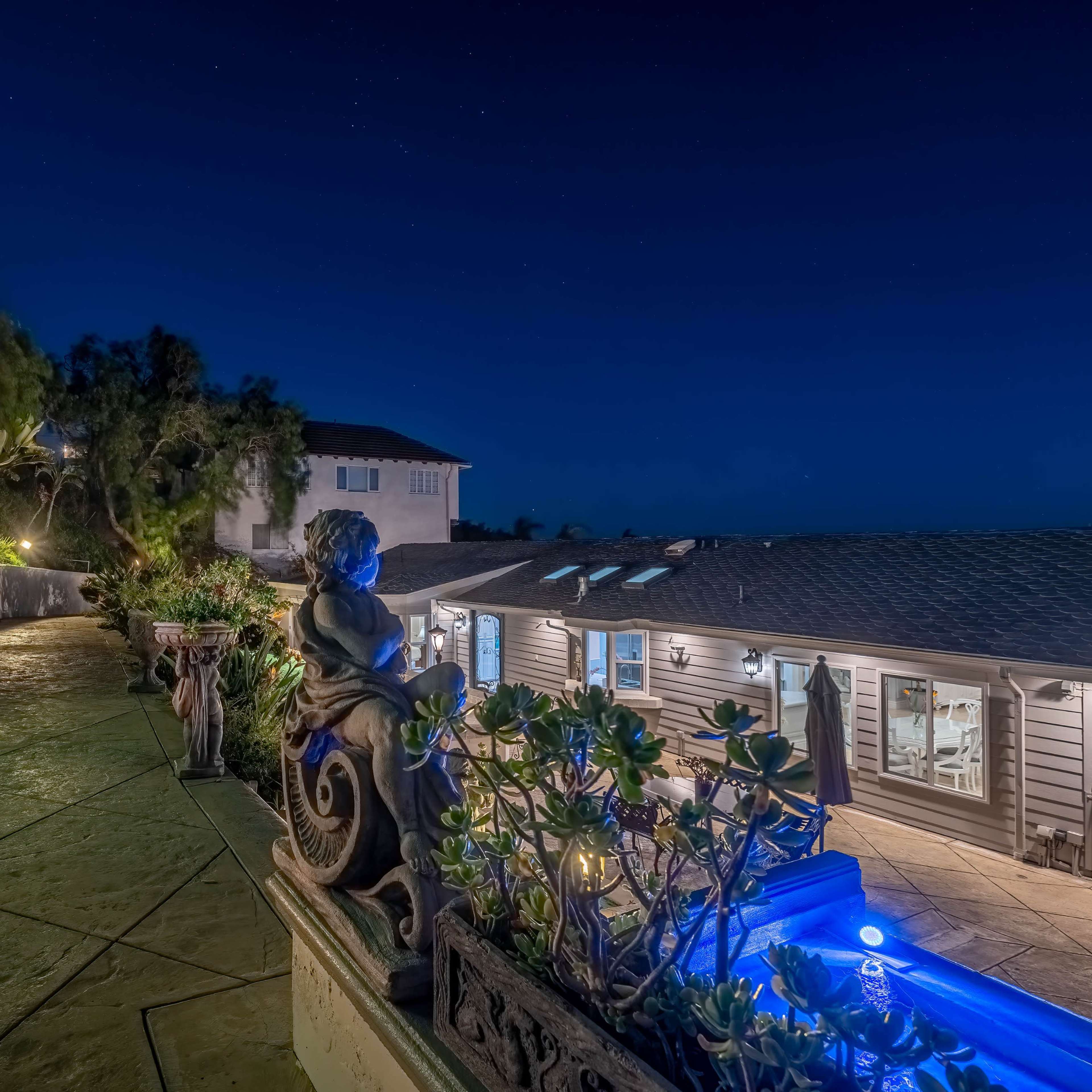 A well-lit outdoor area of a property at night, featuring a stone pathway, a fire pit, and a pool illuminated in blue.
