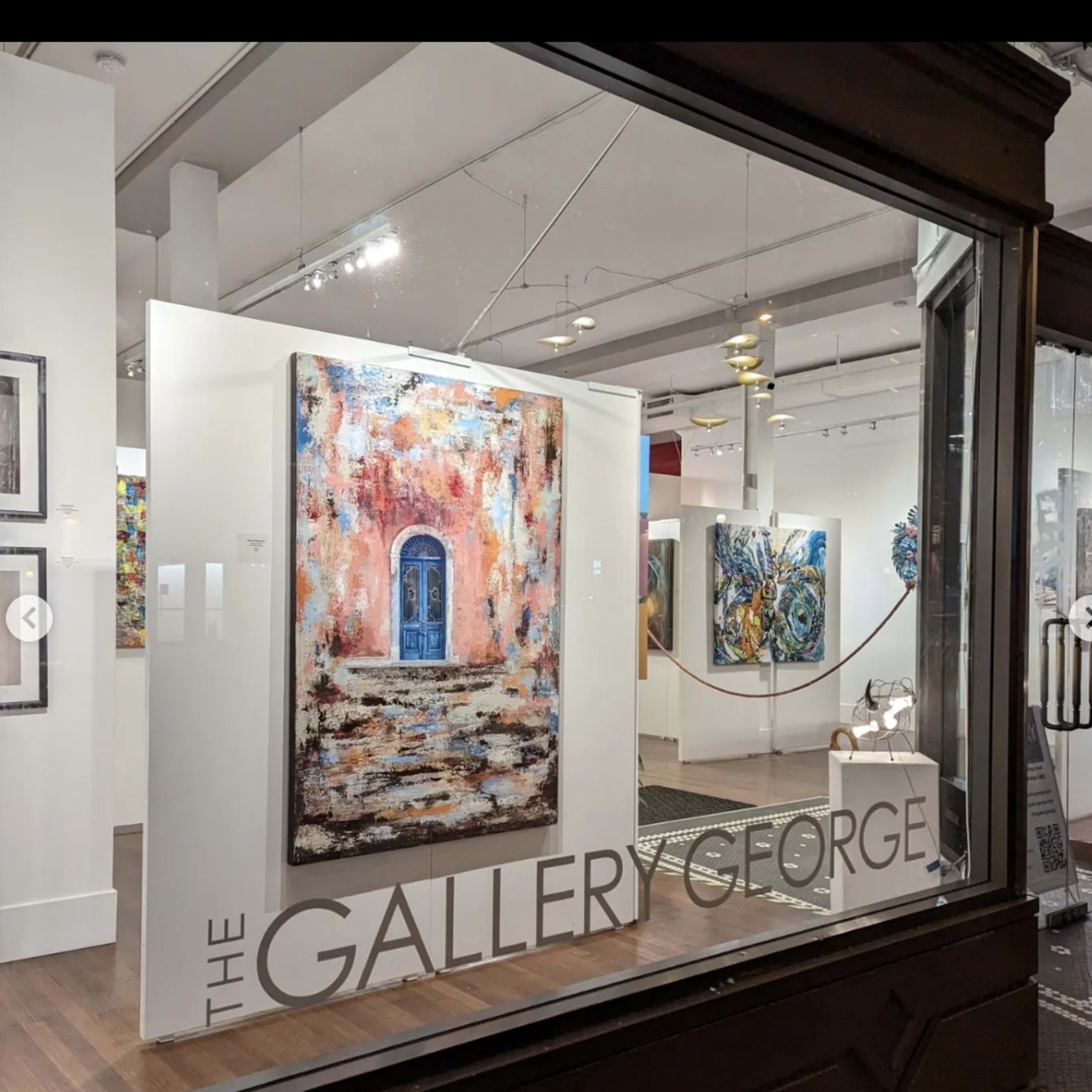 A view of an art gallery's interior, featuring a large painting of a blue door on a pink wall displayed prominently behind a glass window.