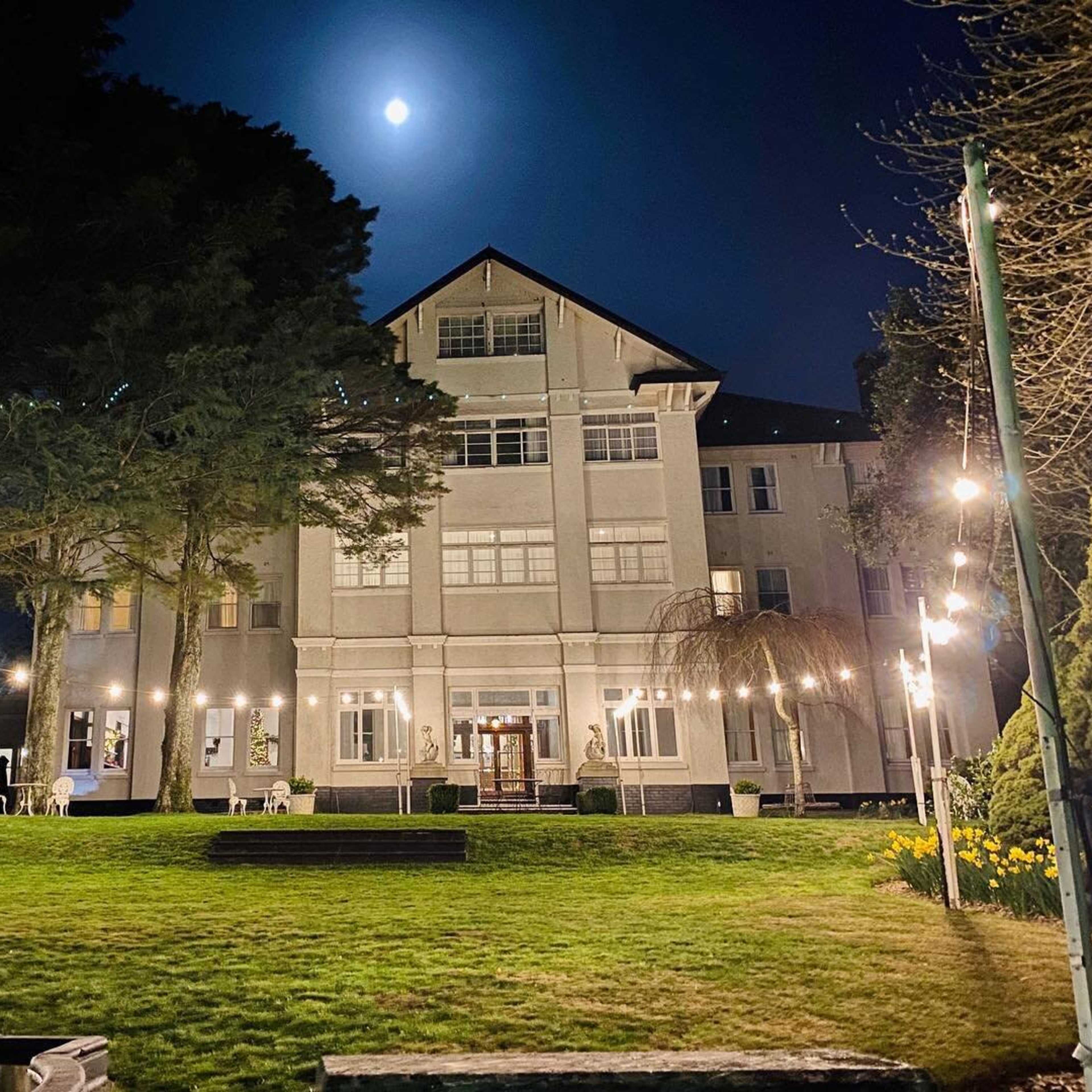 A well-lit, multi-story building is illuminated at night with a full moon overhead and string lights adorning the garden.