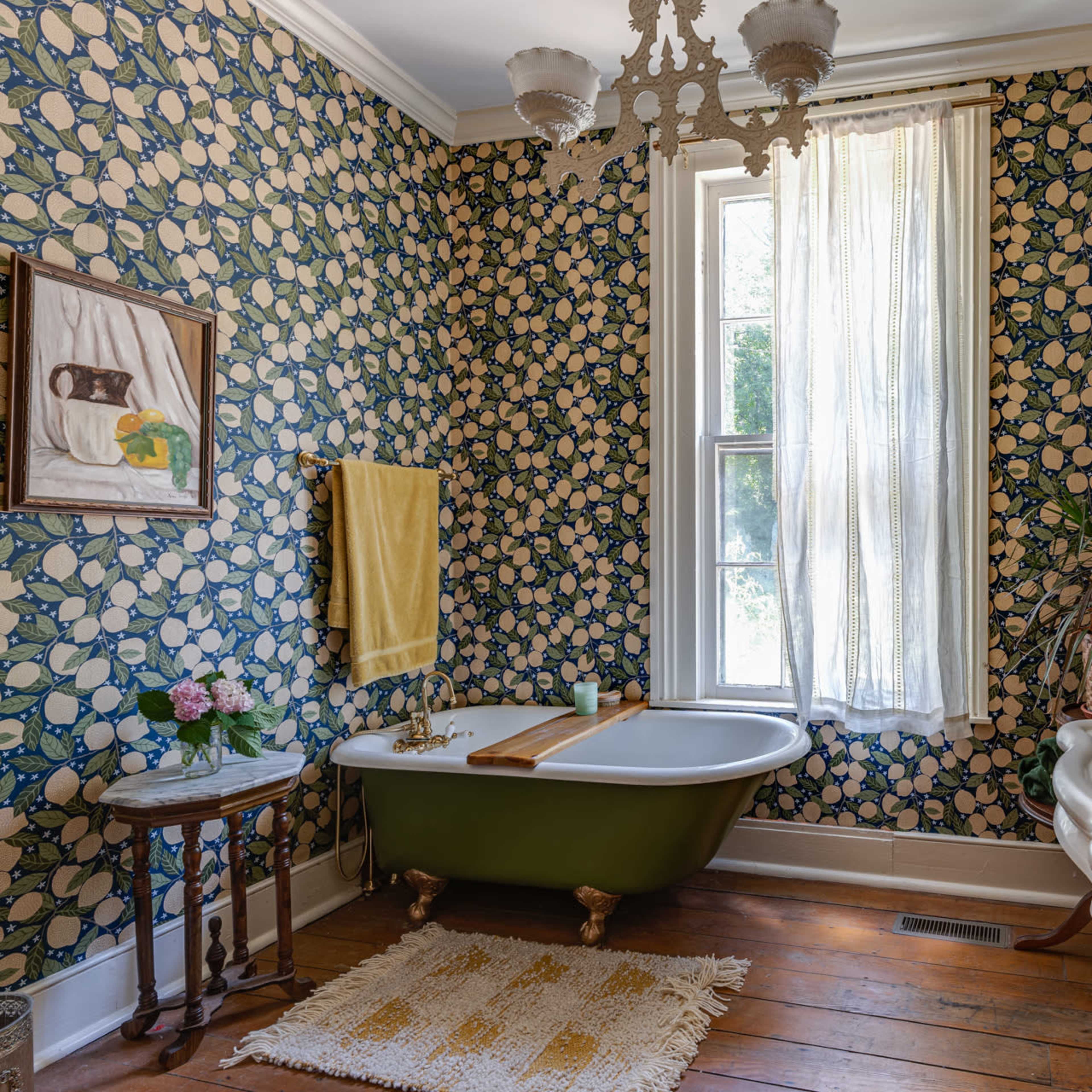A vintage bathroom with floral wallpaper, a green freestanding bathtub, a pedestal sink, and a window allowing natural light to enter.