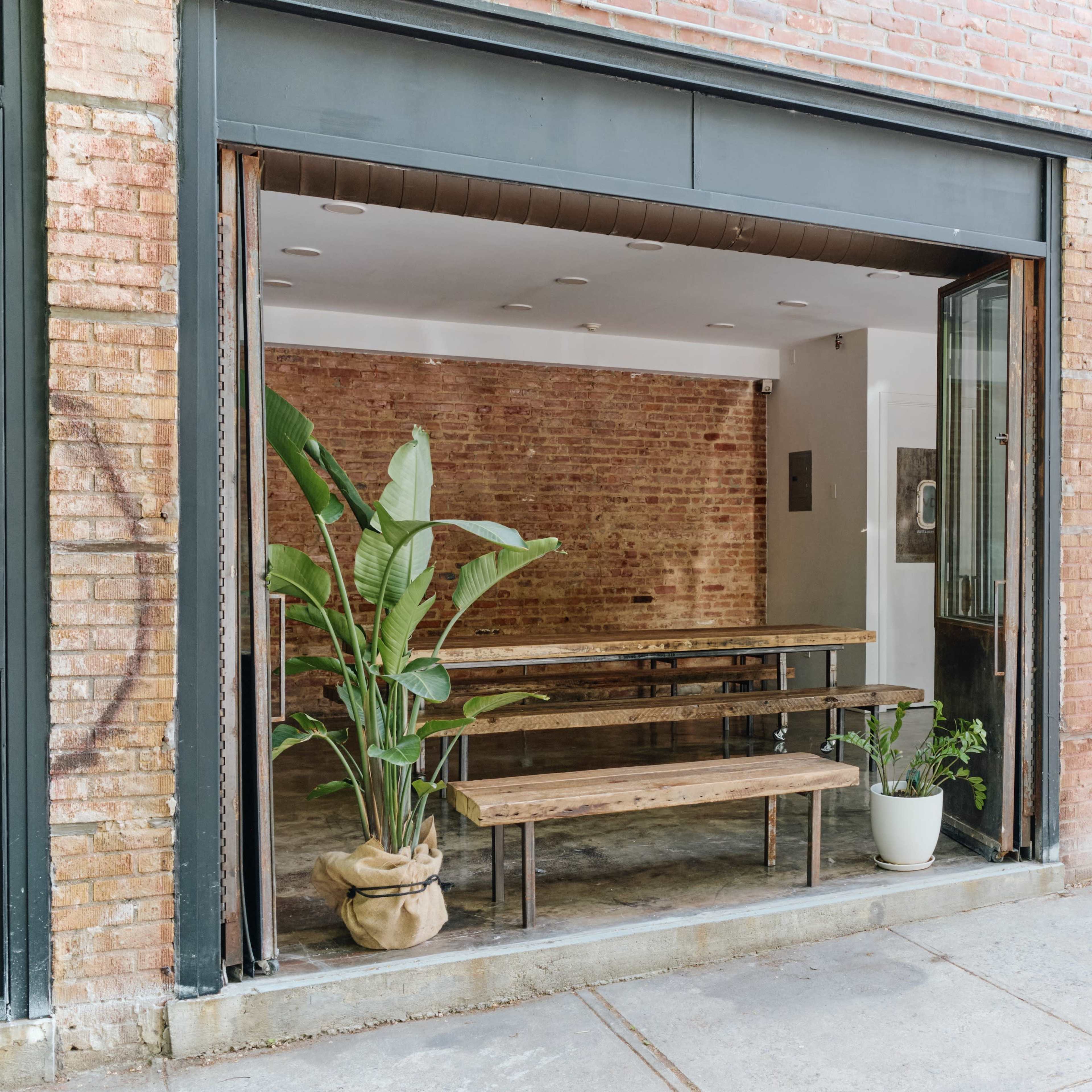 A modern, open storefront with large glass windows revealing an interior featuring brick walls, wooden benches, and potted plants.