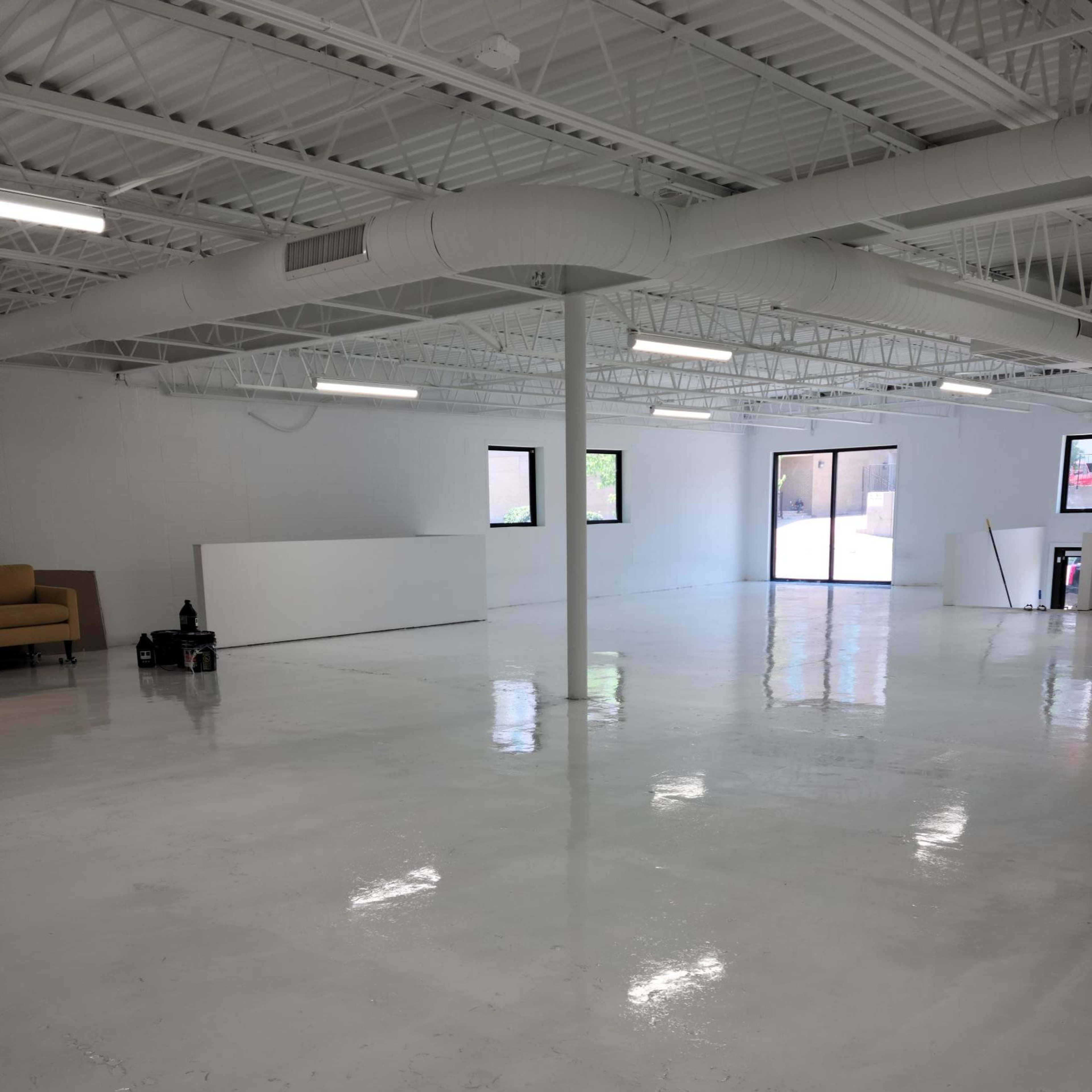 A spacious, empty room with a glossy white floor, white walls, and large windows, featuring minimal furnishings.