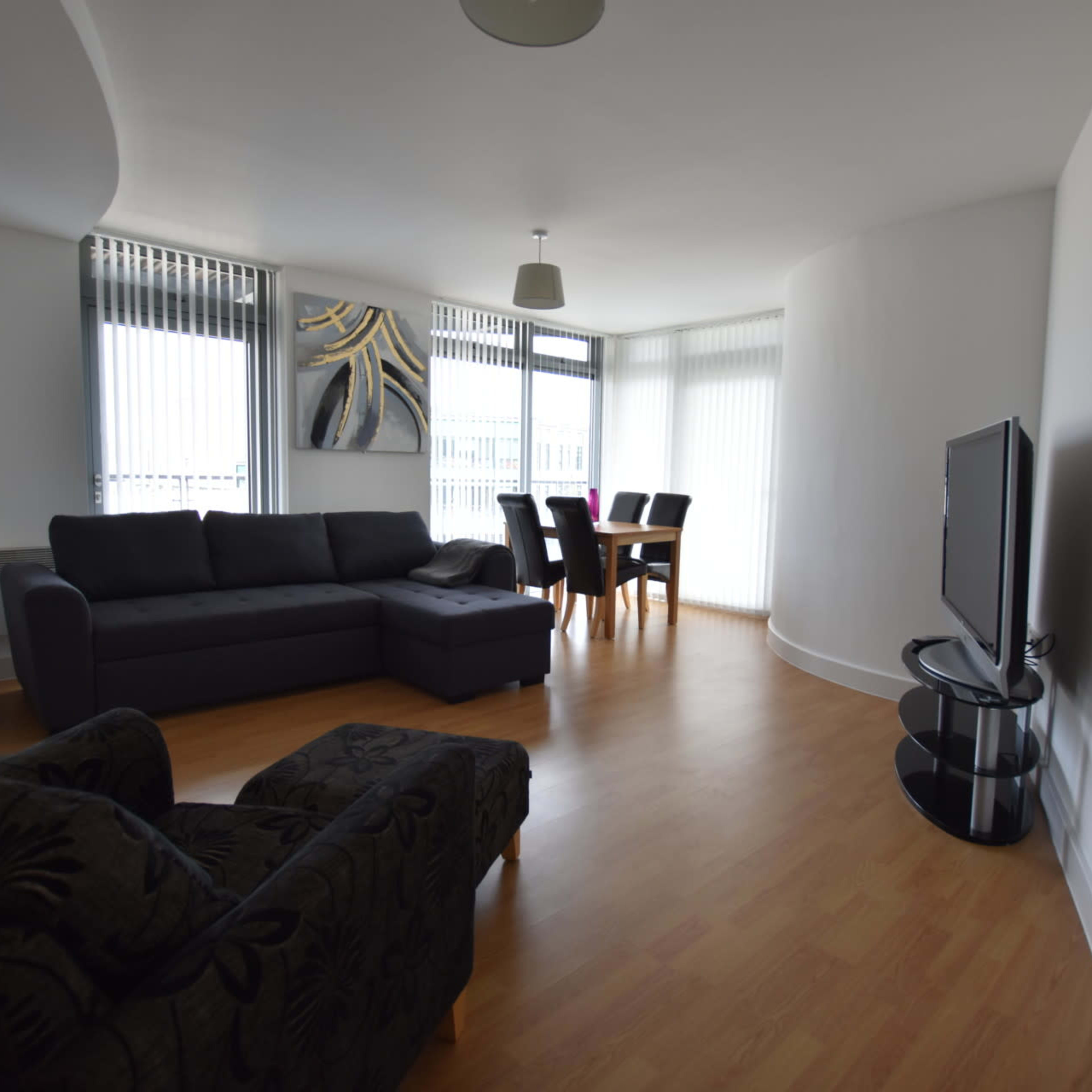 A modern living room with a dark sofa, a black armchair, a dining table with chairs, and a television on a stand, all set against a bright, minimalist interior.