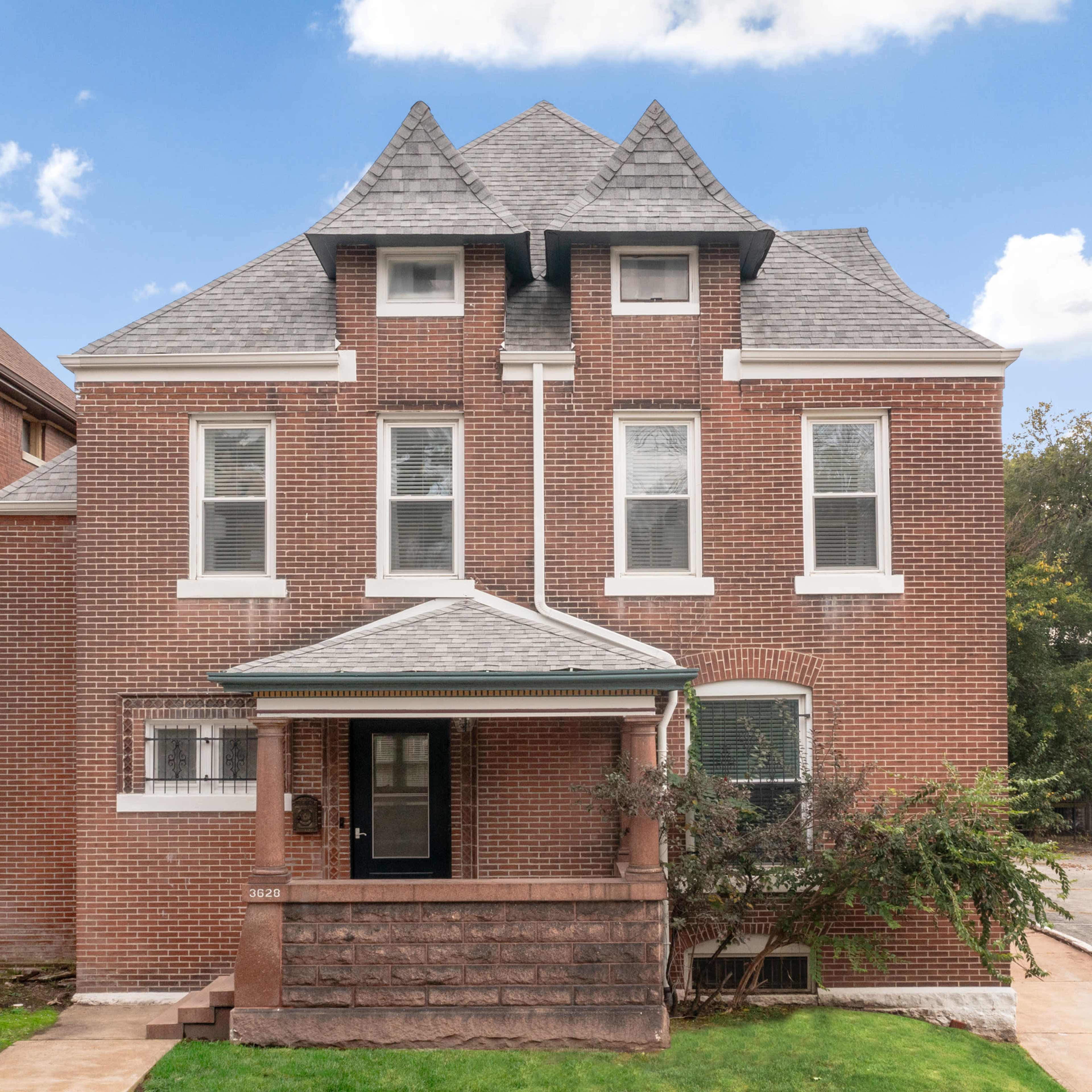 A two-story brick house with a distinctive gabled roof and a porch, situated on a residential street.