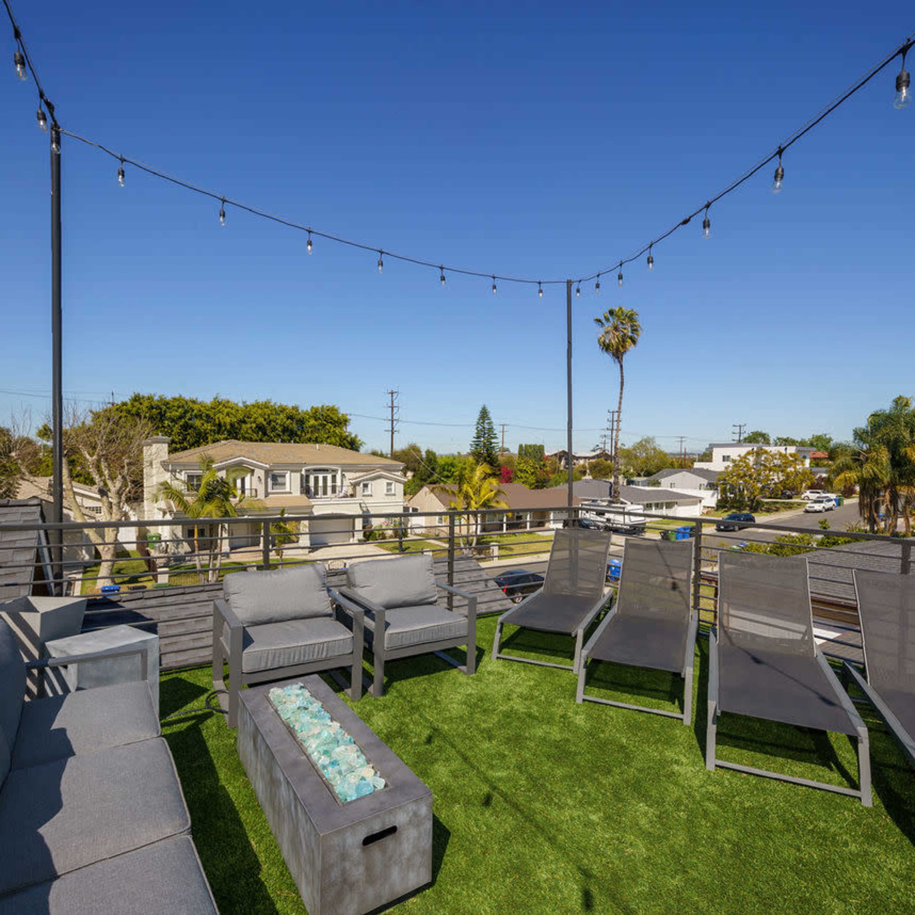 A rooftop patio with gray lounge chairs, a fire pit table, and string lights against a clear blue sky.