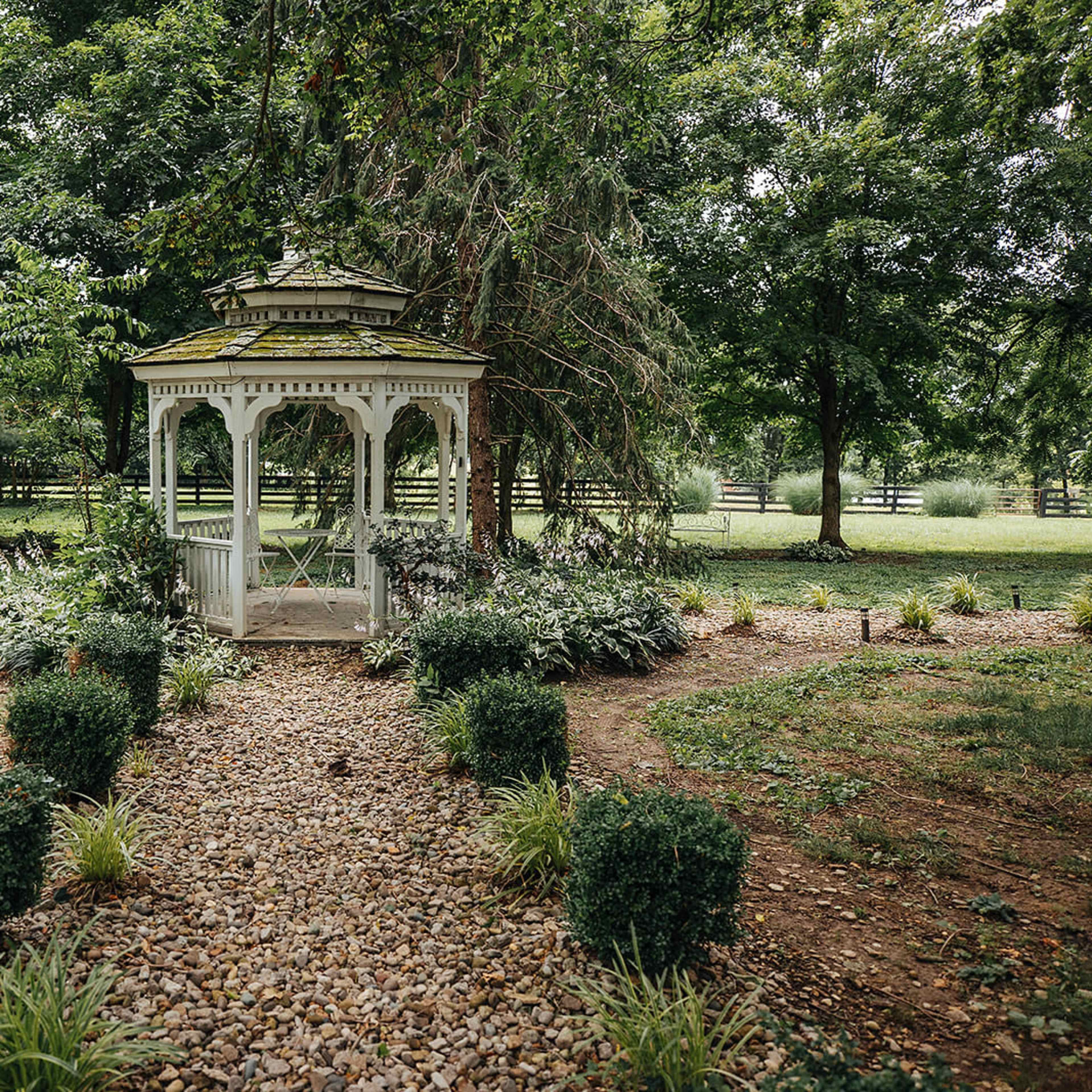 A white gazebo is situated at the center of a landscaped garden surrounded by neatly arranged shrubs and a gravel path.