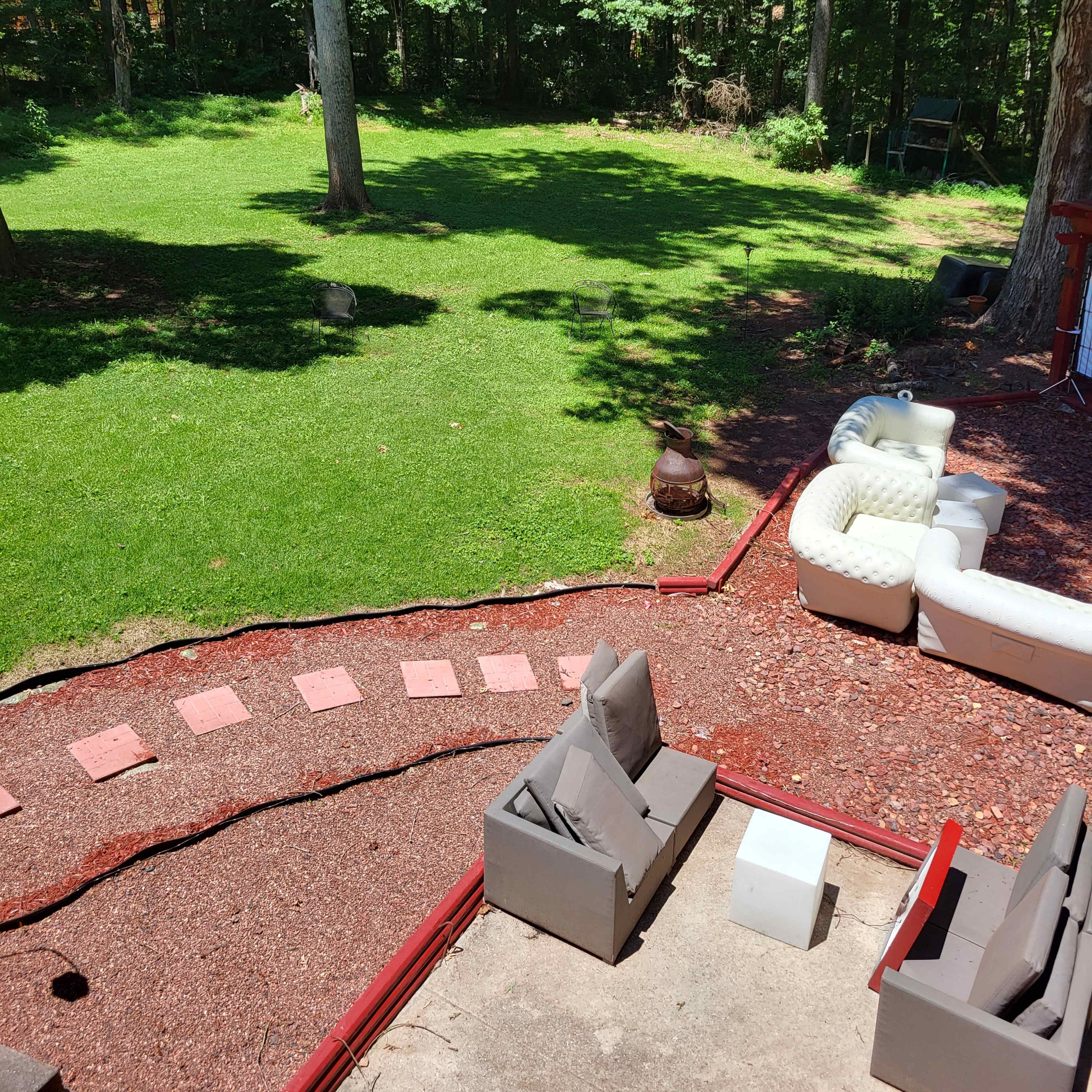 A backyard with green grass, a stone pathway, outdoor seating, and trees in the background.