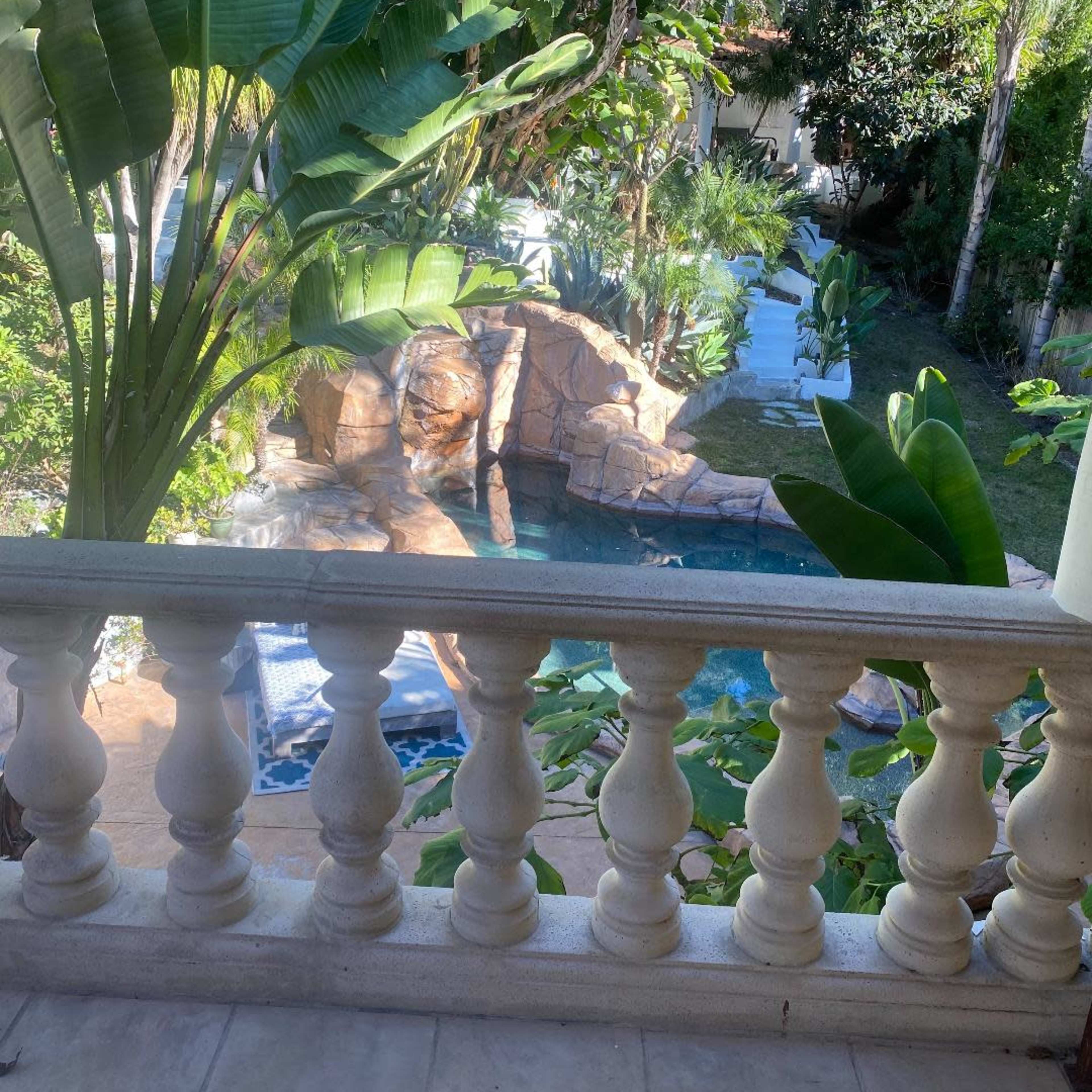 A stone terrace overlooks a swimming pool surrounded by tropical plants and palm trees.