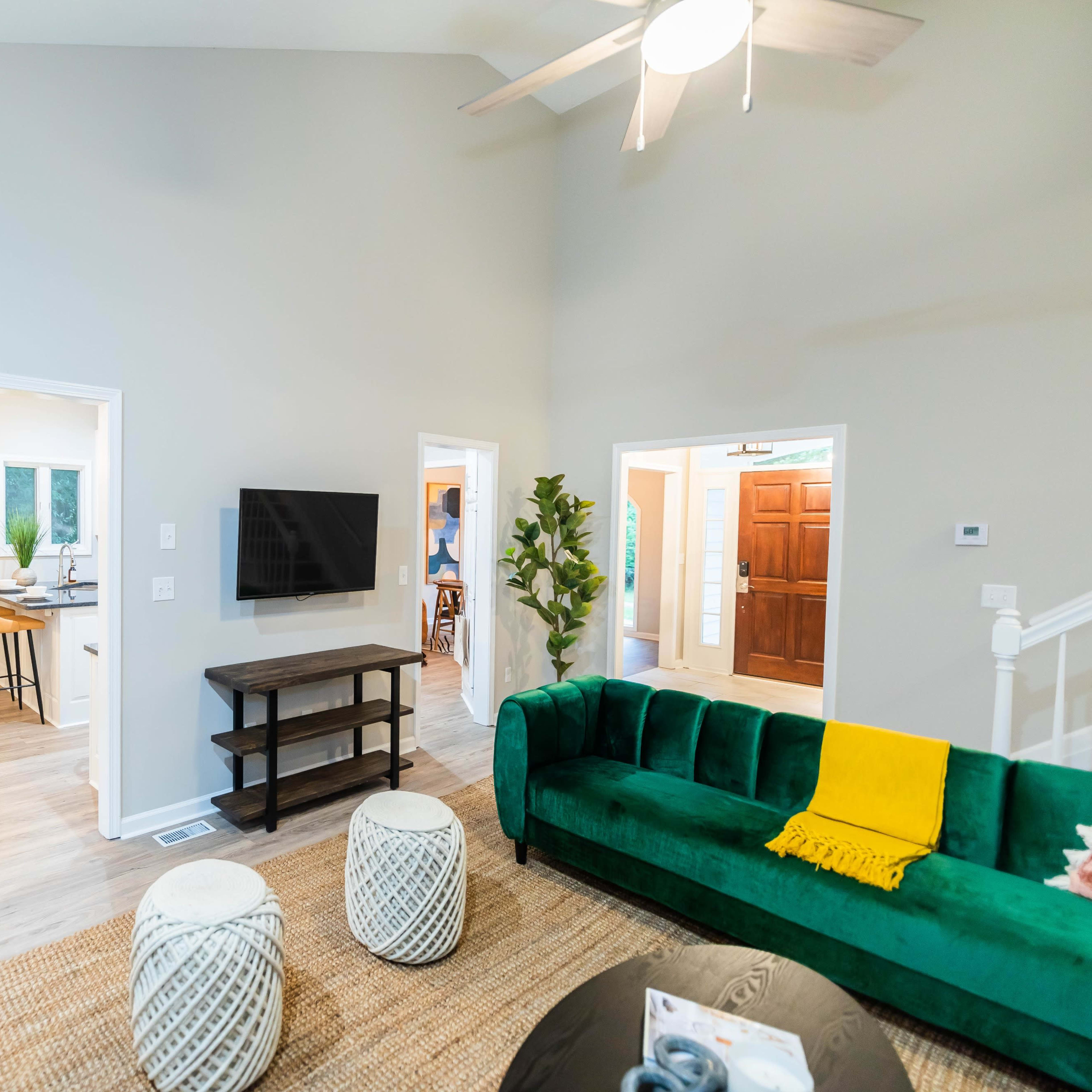 A modern living room features a green couch, a flat-screen TV on a wooden stand, and woven poufs, with open access to adjacent dining and entry areas.