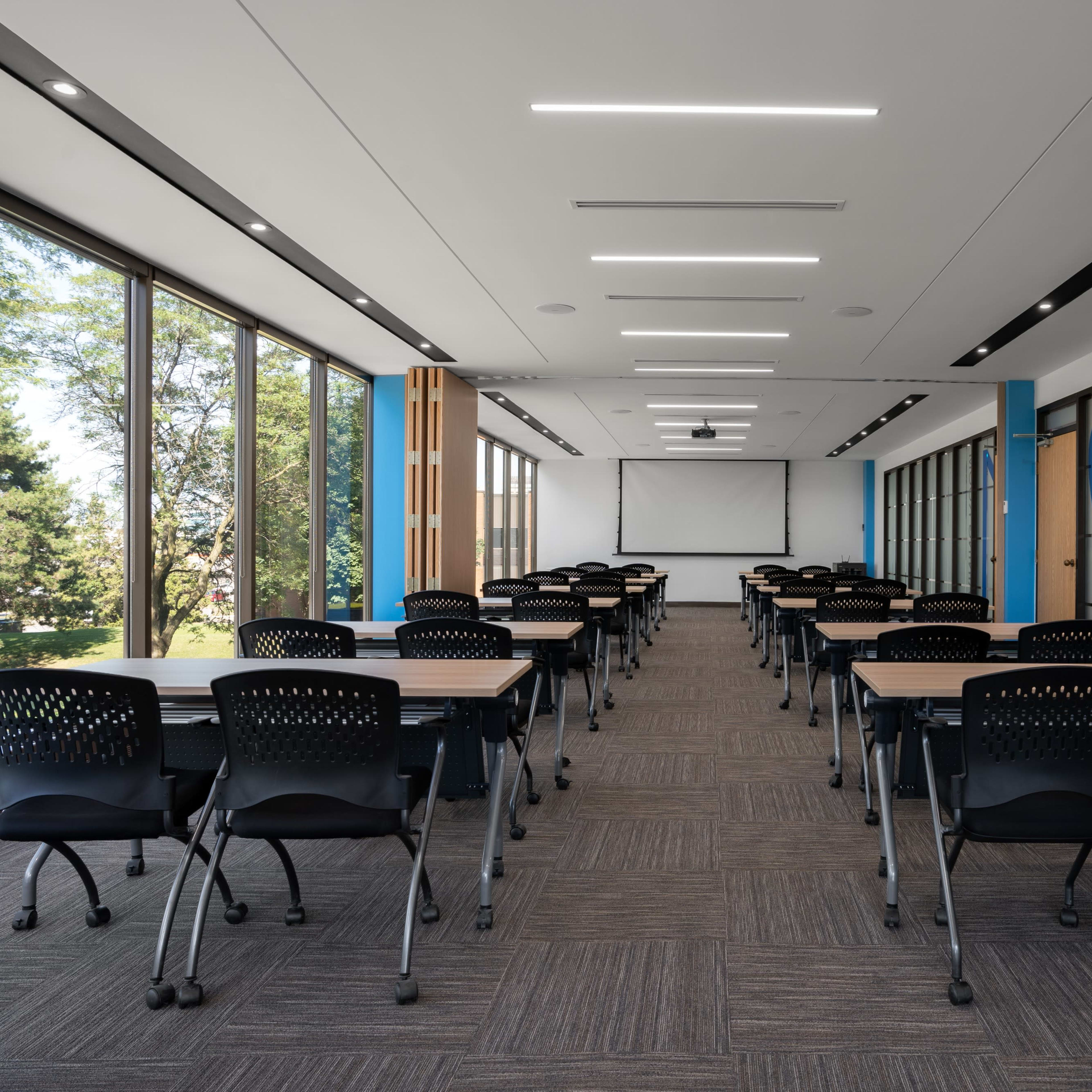 A modern classroom with rows of desks and chairs arranged facing a projection screen, illuminated by natural light from large windows.