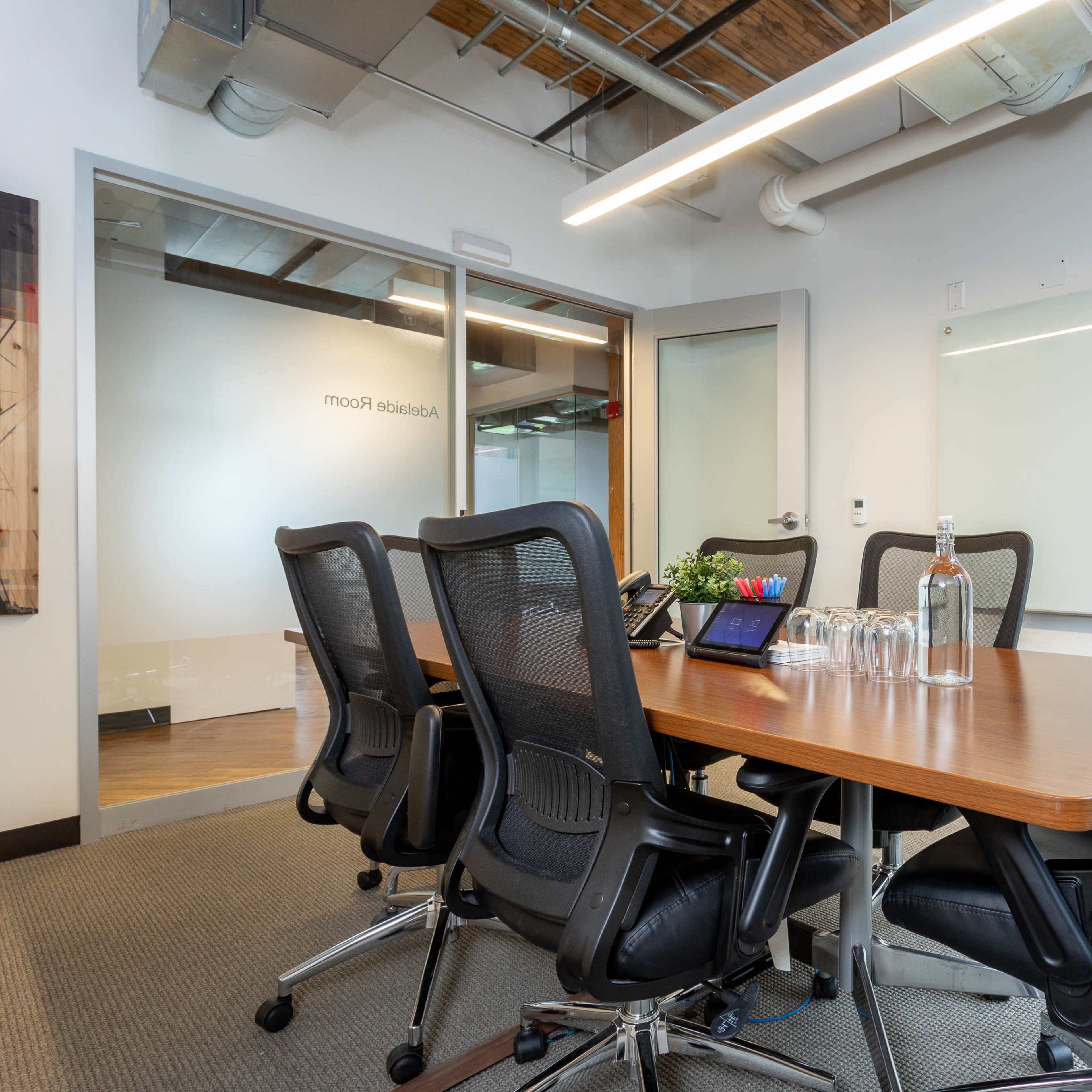 A conference room is set up with a wooden table surrounded by black office chairs, a whiteboard on one wall, and a glass door leading to another room.