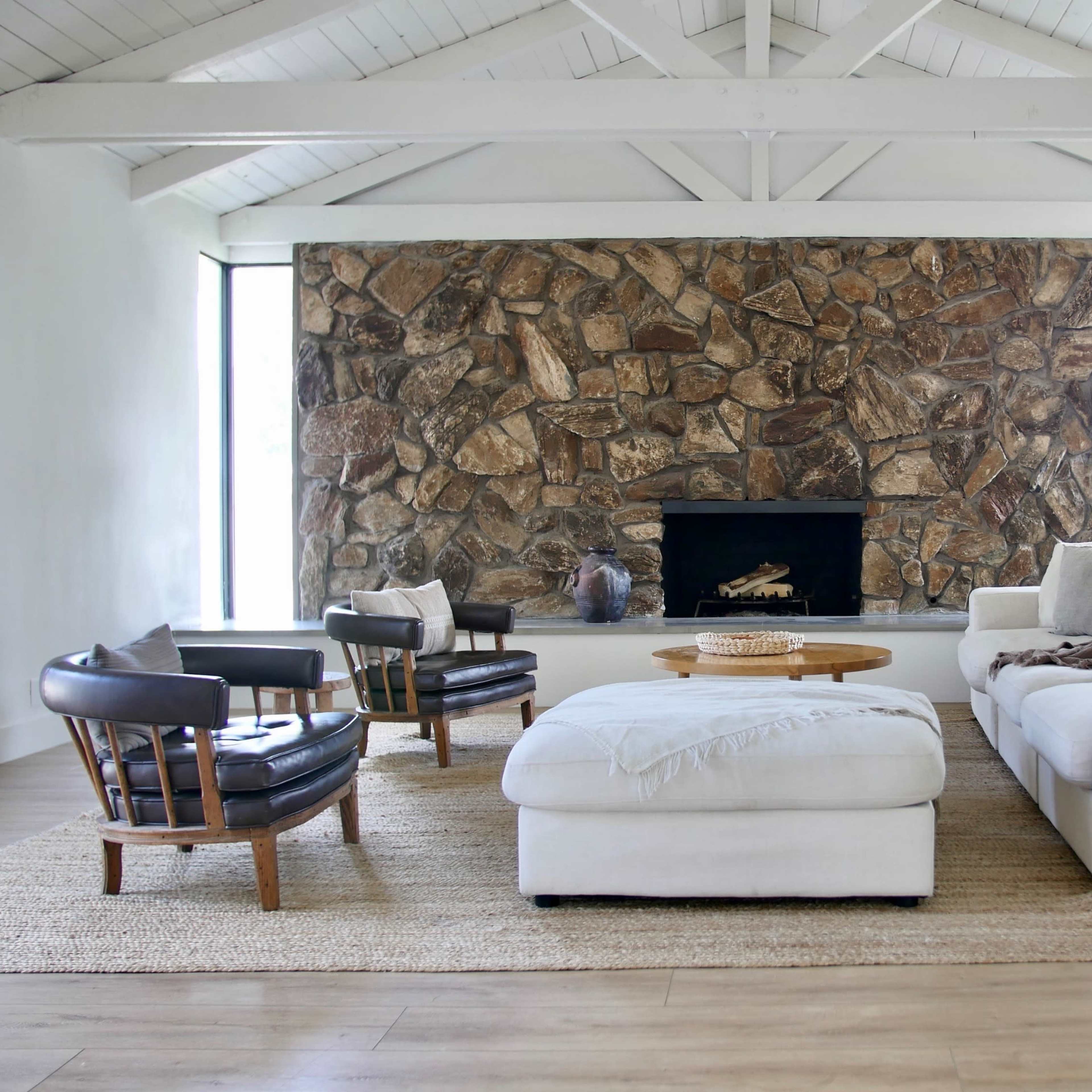 A spacious living room features a large stone fireplace, a white sofa, a round coffee table, and several armchairs arranged around a woven rug.