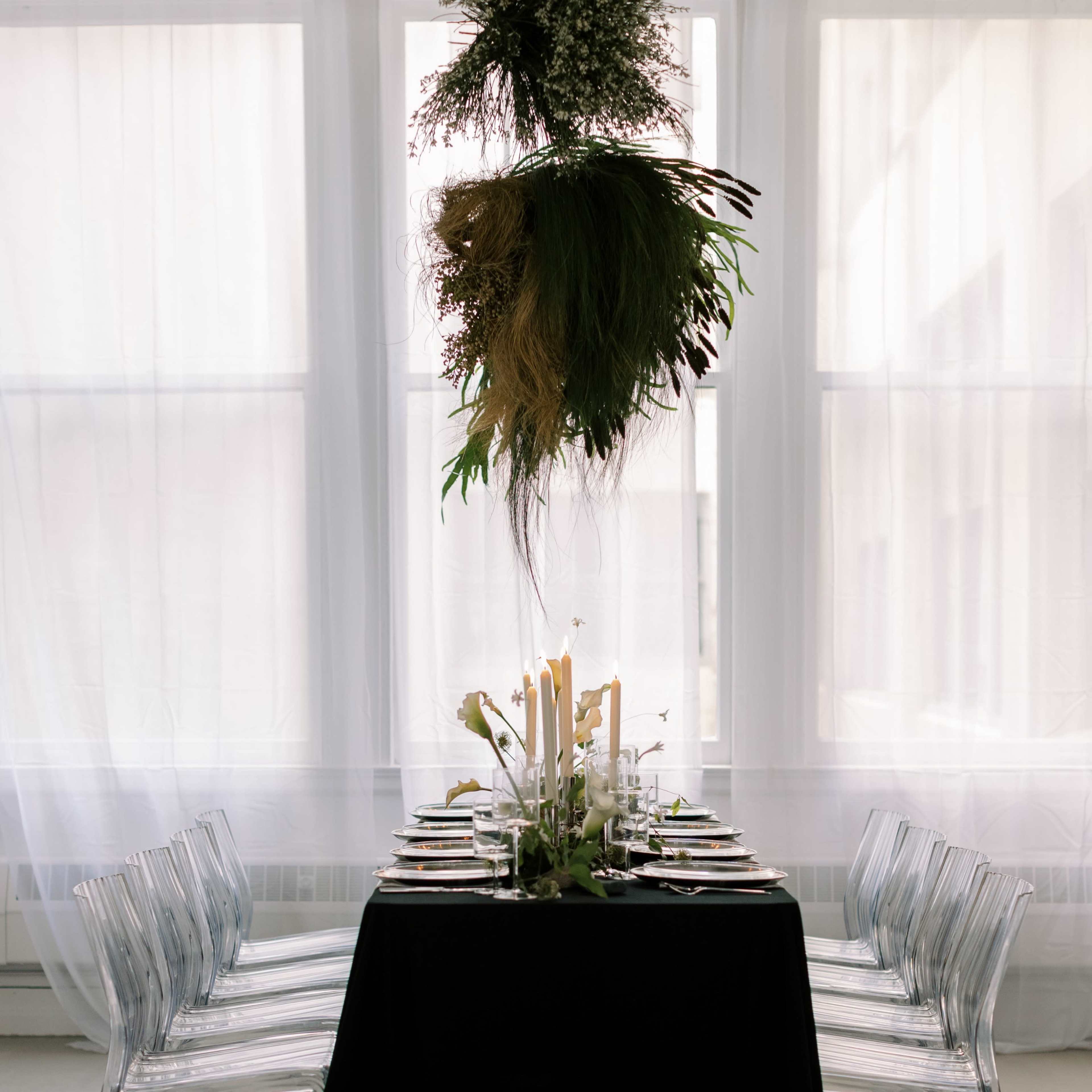 A dining table is set with elegant tableware and surrounded by transparent chairs, with a floral arrangement hanging above in a well-lit room.