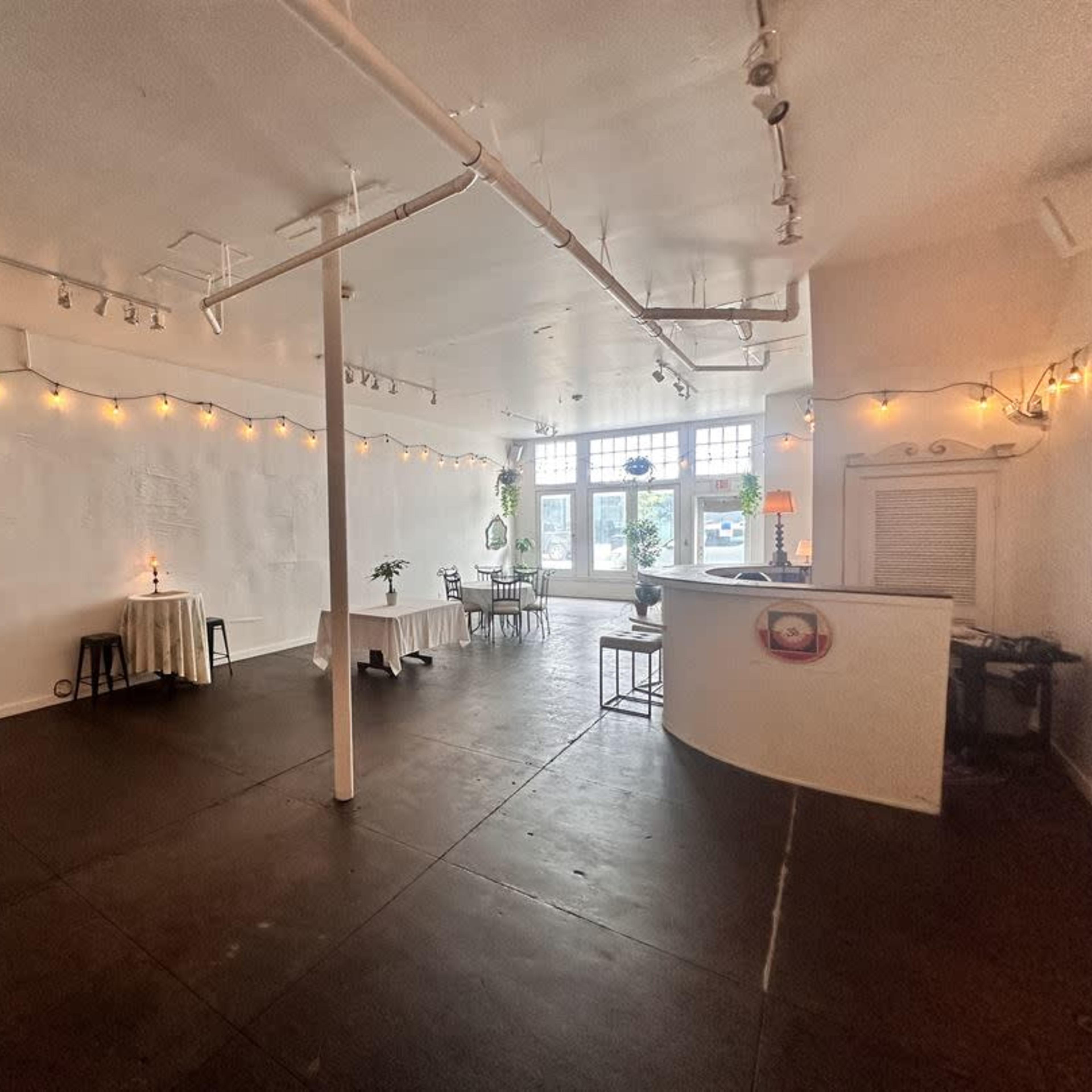 An empty, well-lit interior space with white walls, a dark floor, and a few tables near the windows.