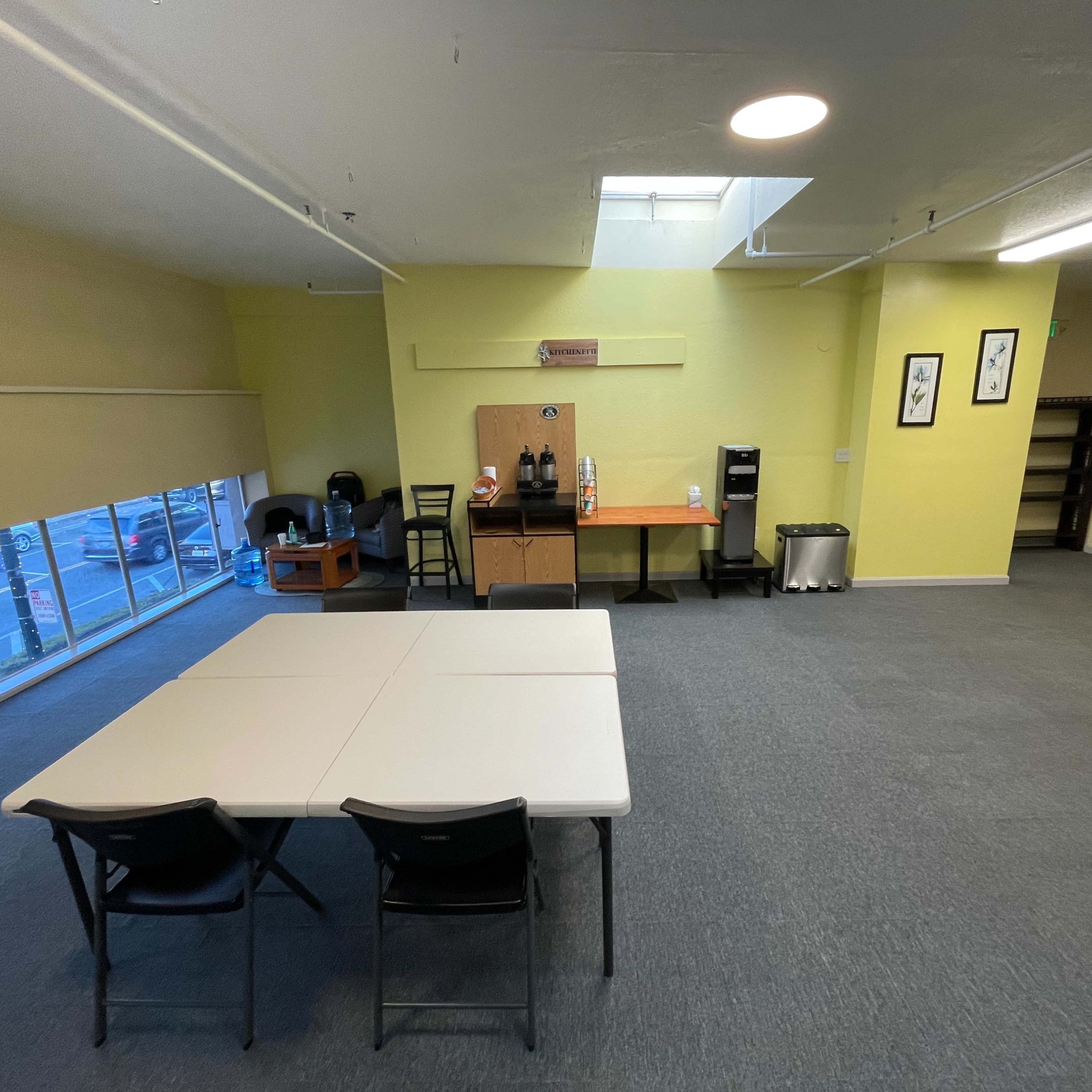 A spacious, brightly lit room featuring a central table with chairs, a coffee station, and large windows overlooking a parking area.