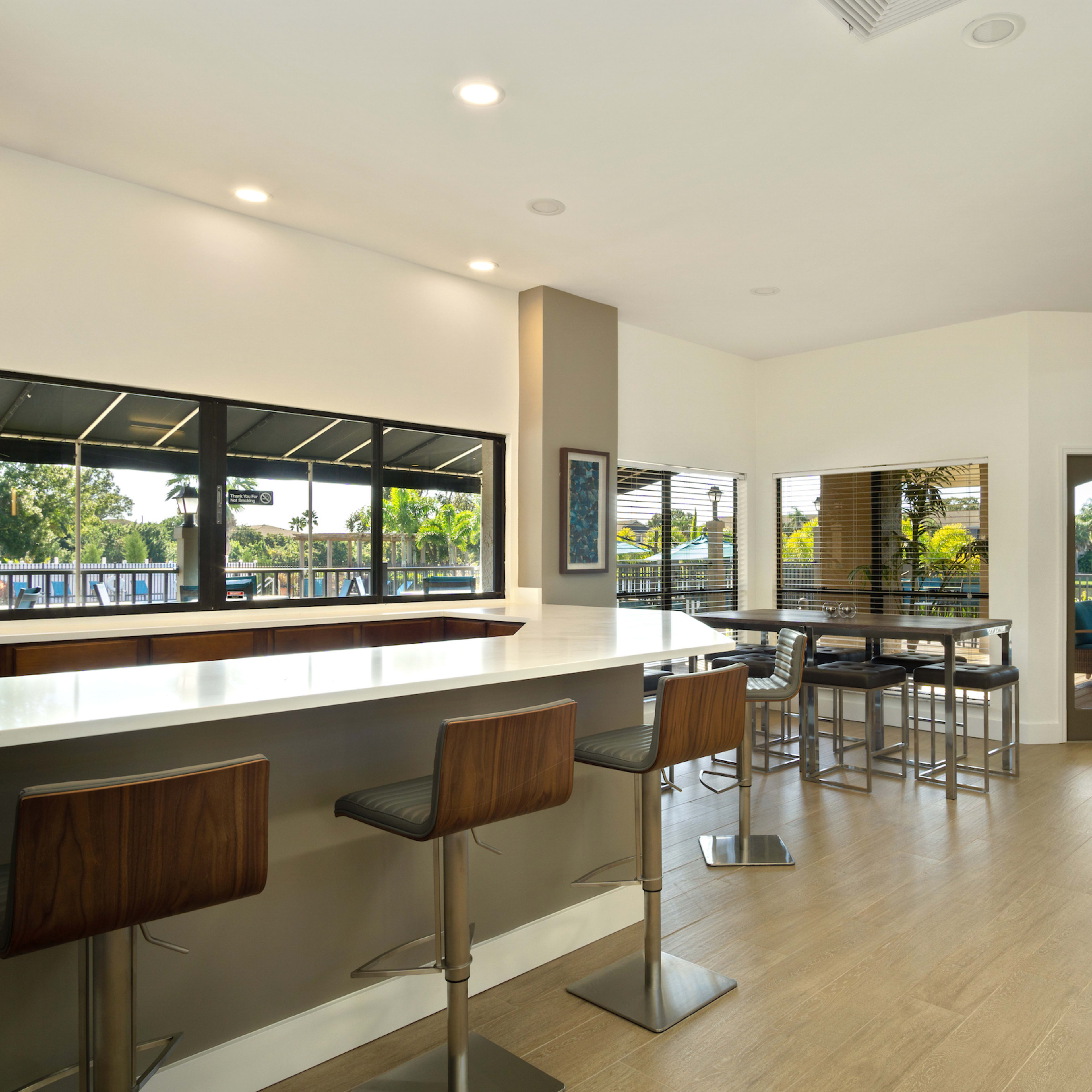 A modern interior space with a long countertop, bar stools, large windows overlooking greenery, and a seating area with additional tables and chairs.