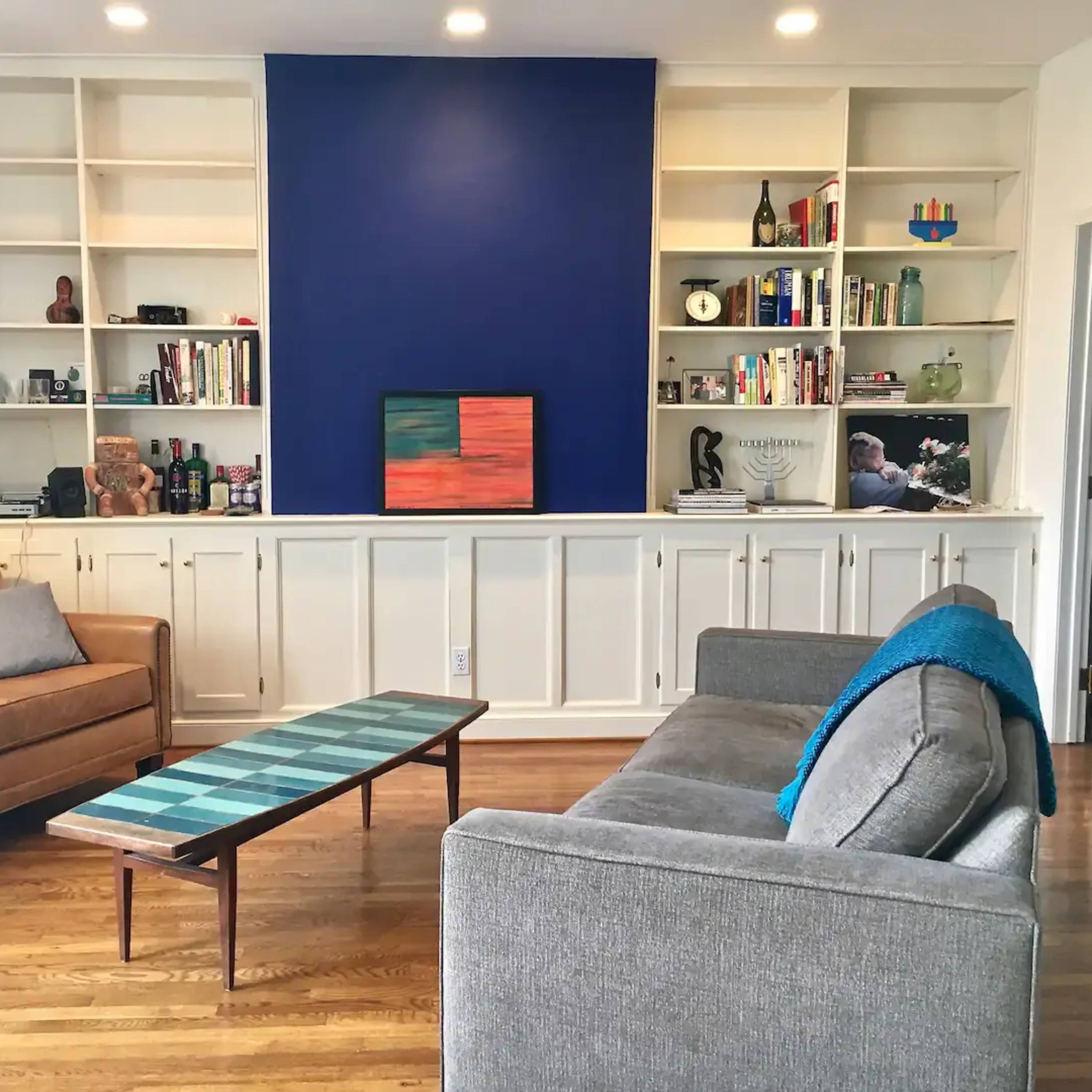 A living room with two sofas facing a low coffee table, a bookshelf filled with various items, and a blue accent wall.