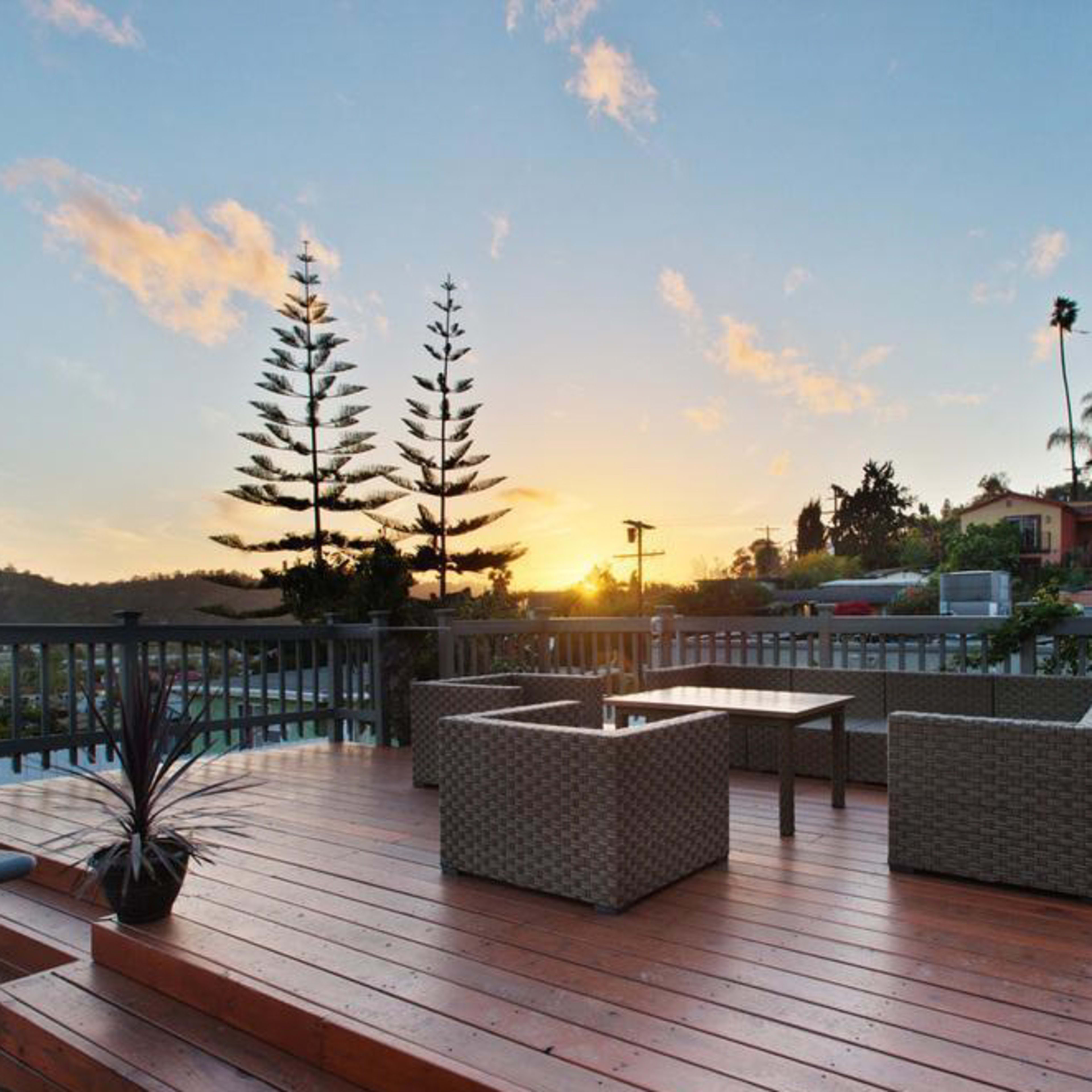 A wooden deck features two chairs and a table, set against a backdrop of a sunset with palm trees and houses in the distance.