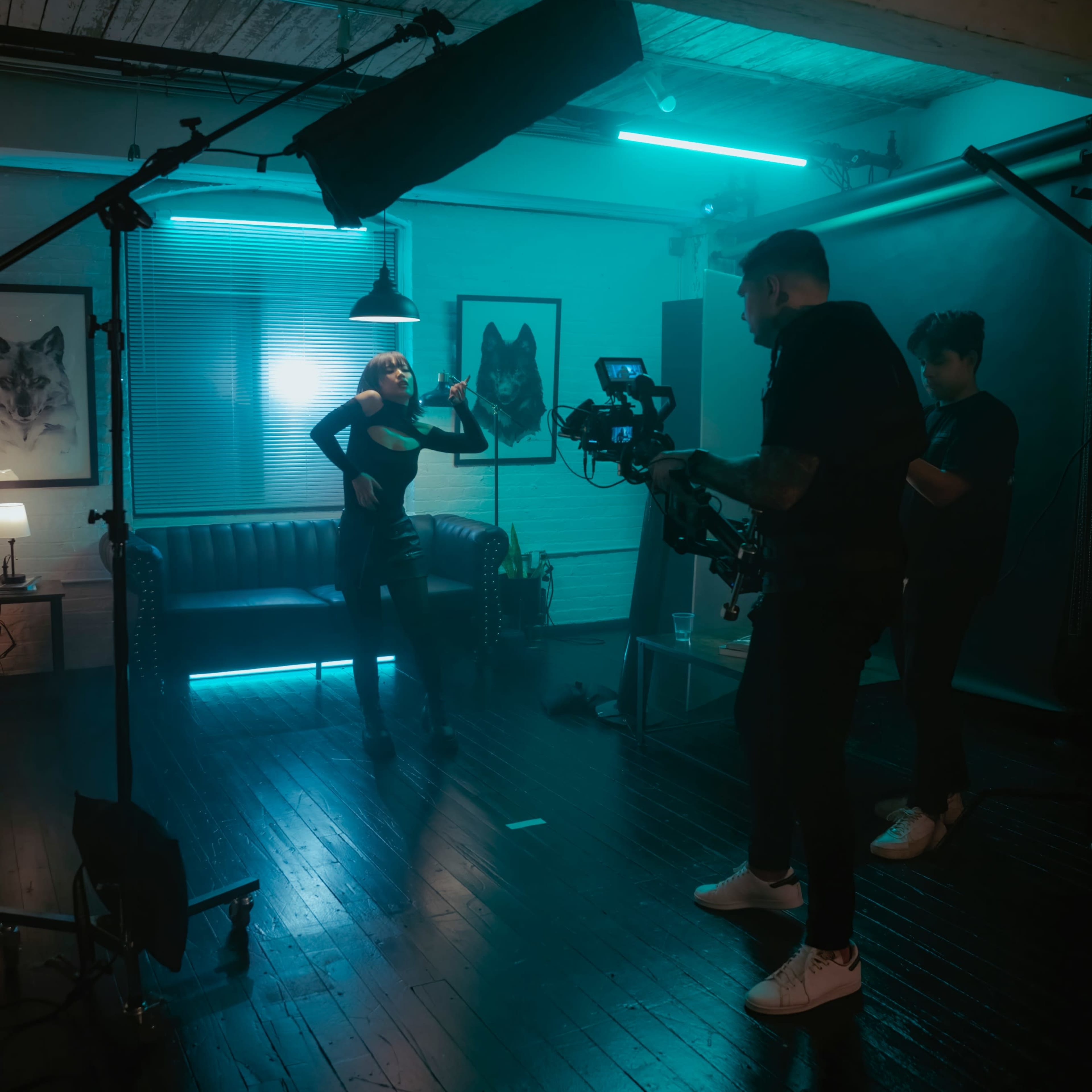 A photoshoot takes place in a dimly lit room featuring a model posing near a couch, while two crew members operate a camera and lighting equipment.